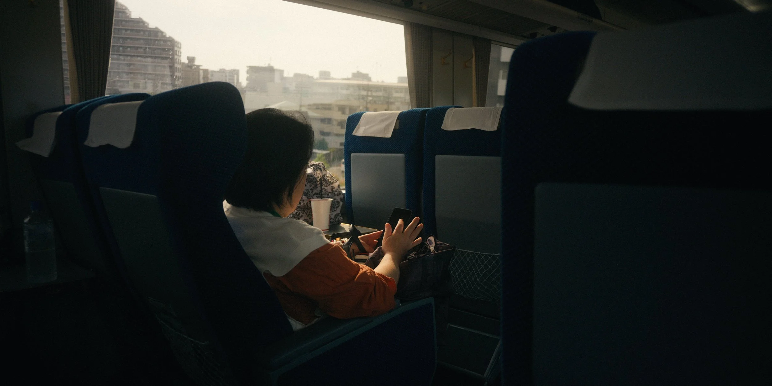 Inside a bus, a woman is sitting and using her phone. The scene shows the backs of bus seats with headrests, a water bottle on the window ledge, and city buildings visible through the large front window, under overcast or cloudy weather.