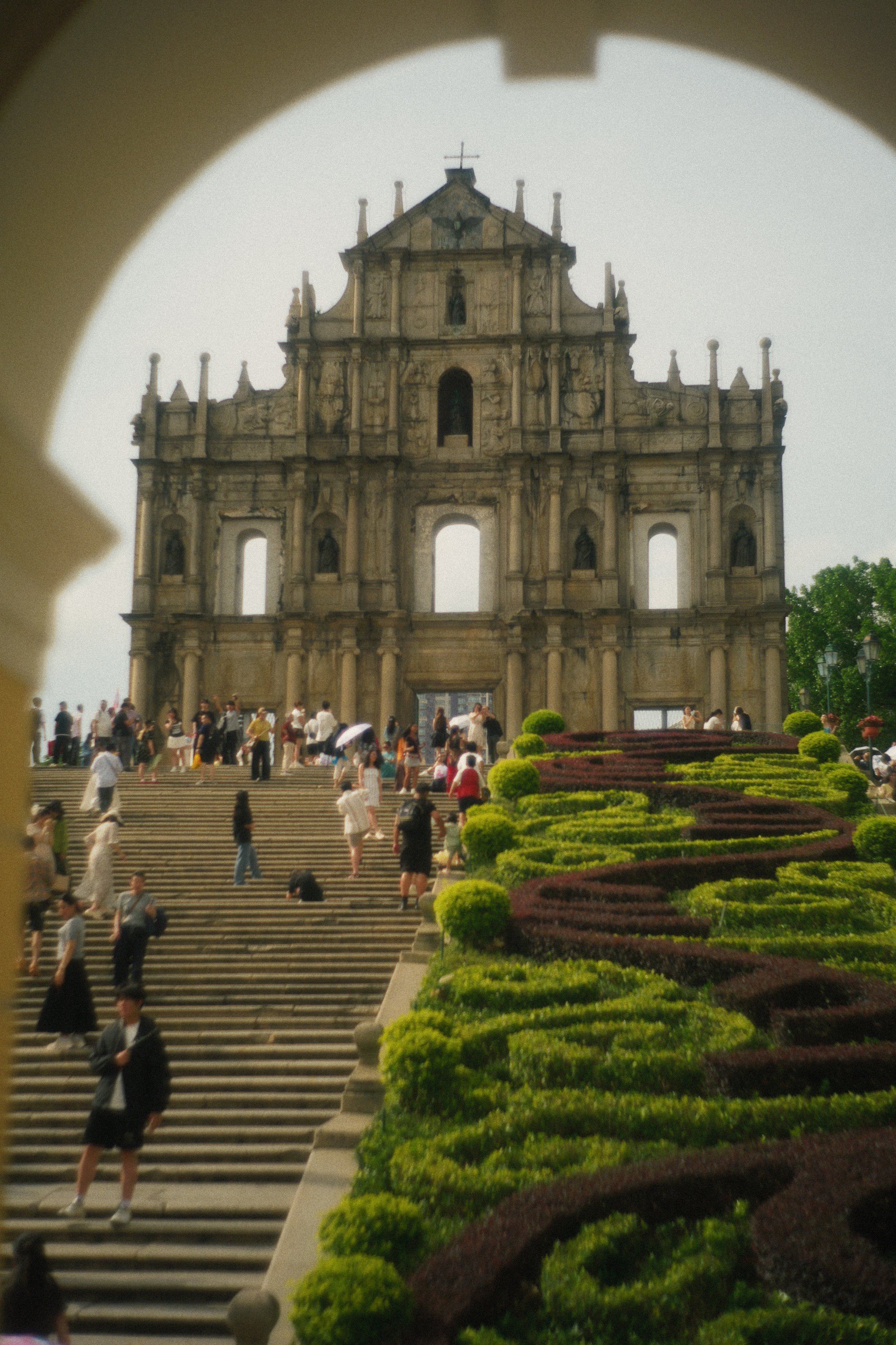 A historic stone archway with multiple statues and decorative elements on top, viewed through a monument or frame. People are walking up and down stairs leading to the archway, and there are well-maintained gardens with green and dark red bushes in t
