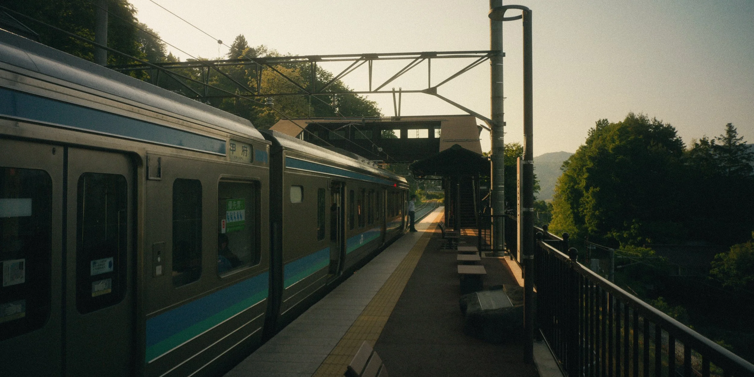 A train at an outdoor station surrounded by trees during sunset, with a person waiting on the platform.
