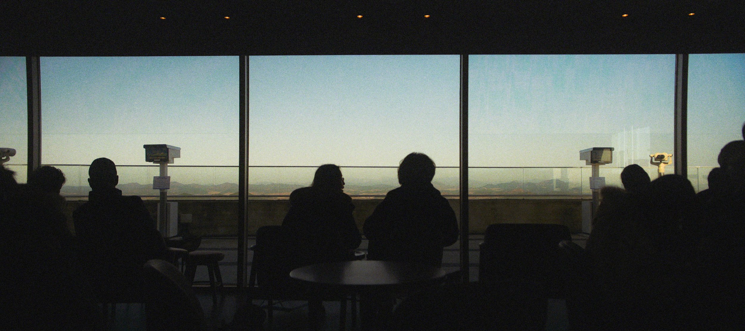 People sitting in a dark room at an airport terminal, looking through large windows at a clear sky and distant mountains.