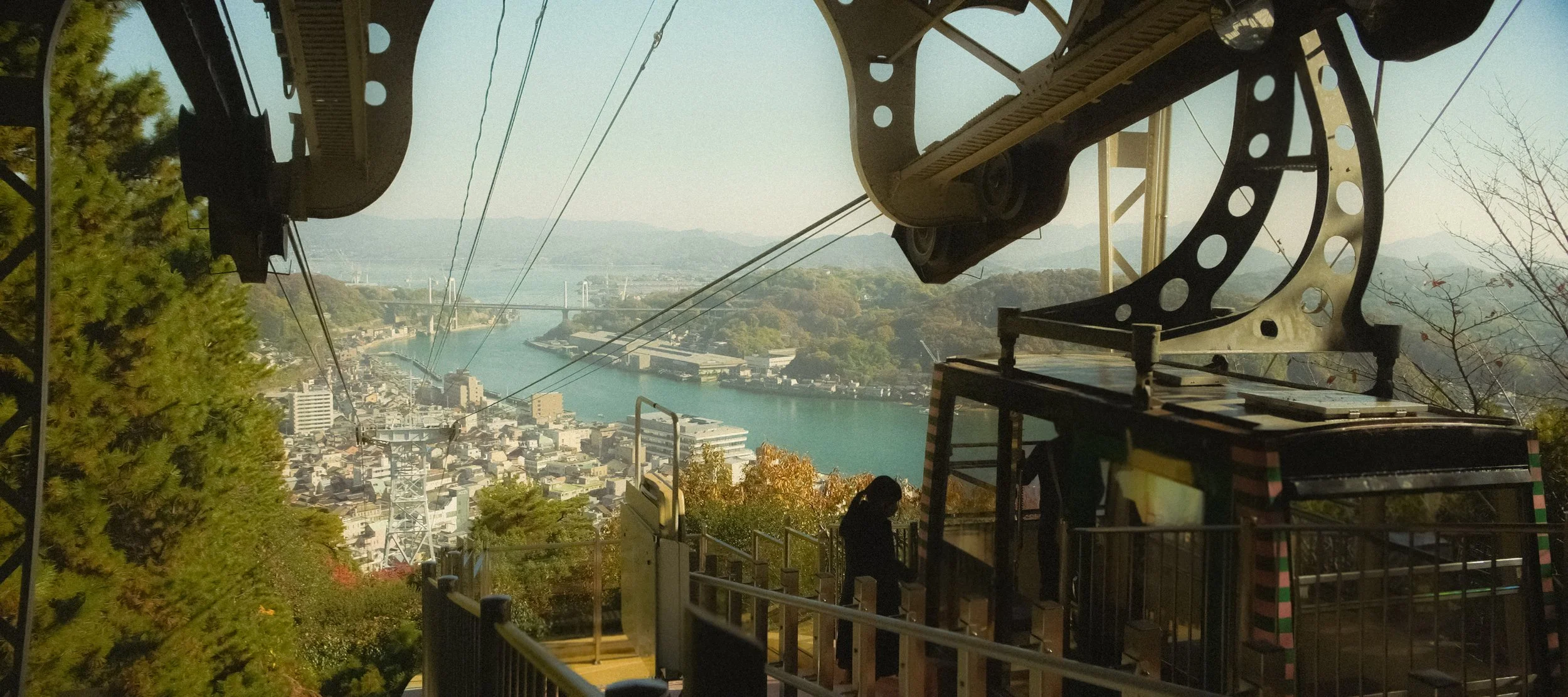 View from a cable car showing cityscape, river, and mountains in the background, with mechanical equipment and trees in the foreground.