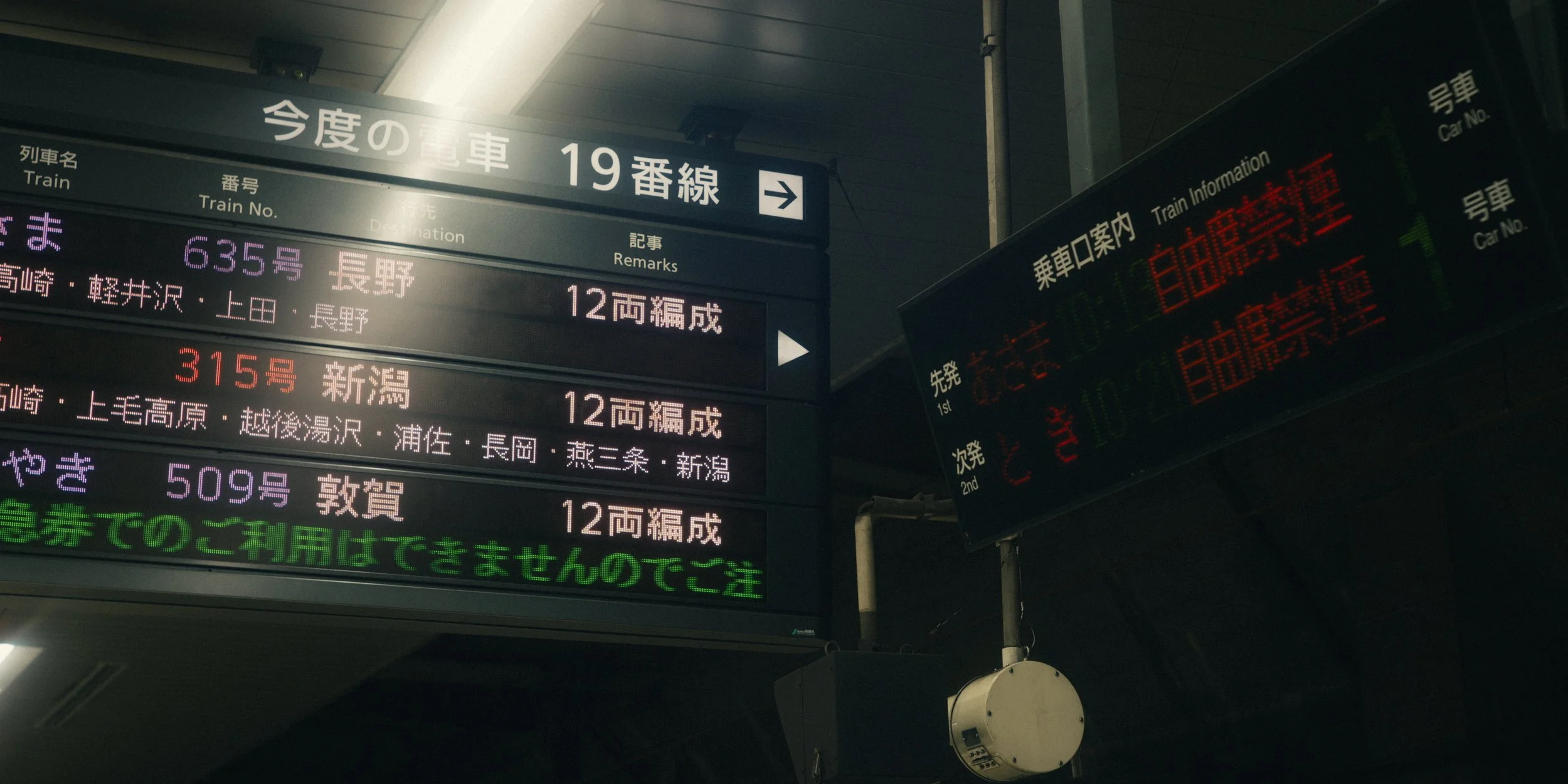 Electronic train information displays showing train routes and schedules in Japanese, with some text in English, in a train station.