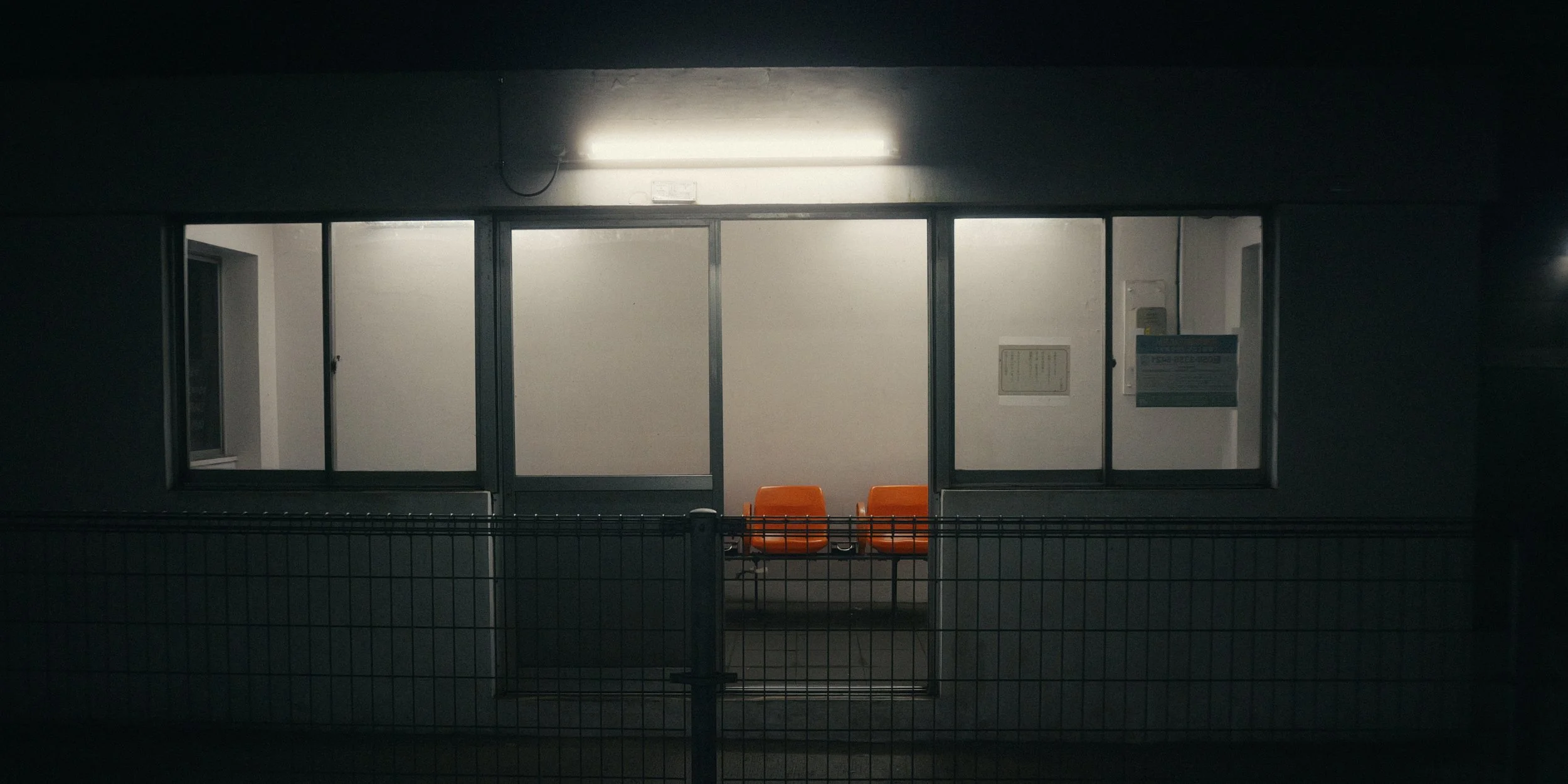 Night view of a bus stop with orange seats, illuminated from above, surrounded by a black fence.