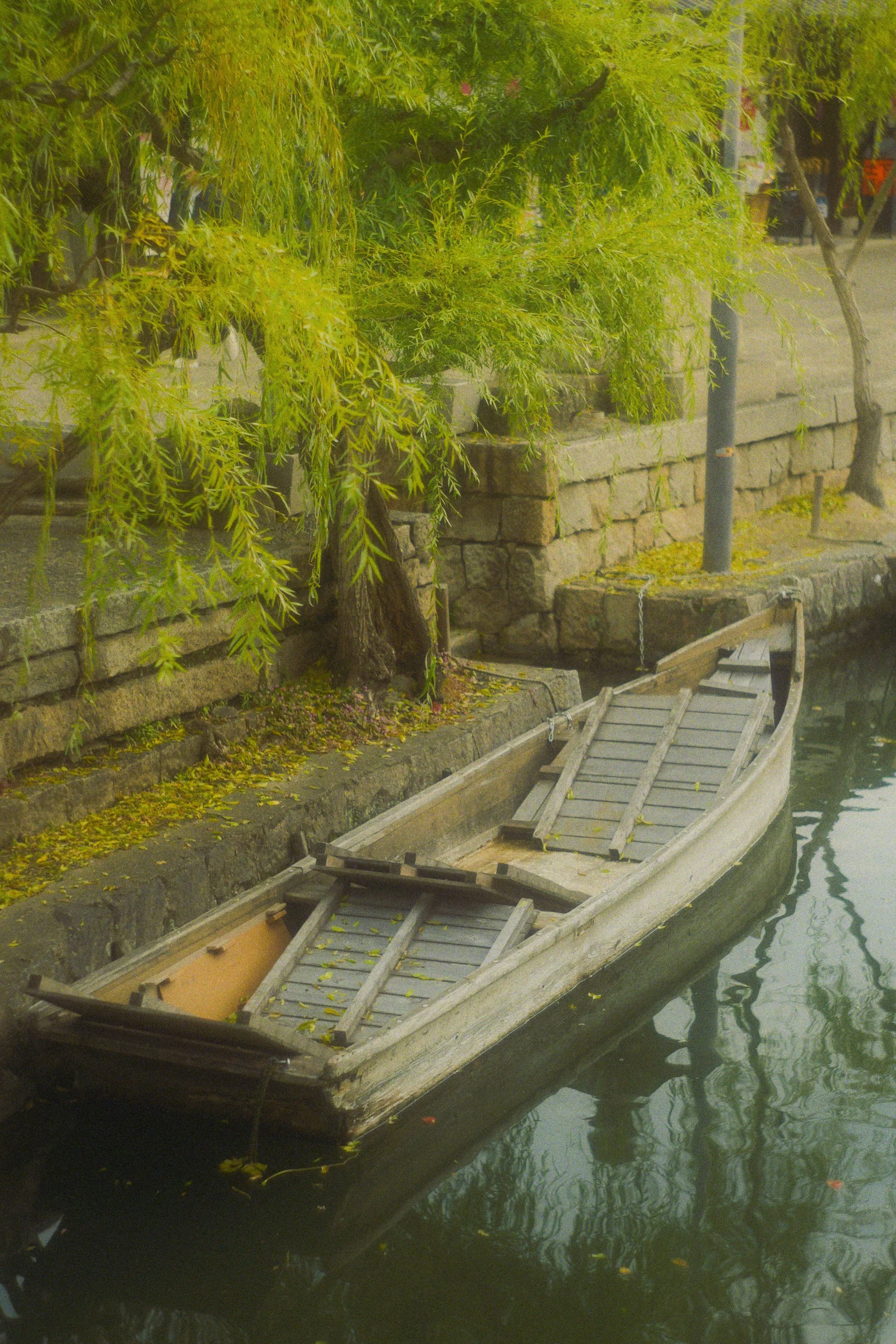 A small wooden boat floating in a canal with a stone wall and green trees in the background. The boat appears to be empty with some fallen leaves inside.