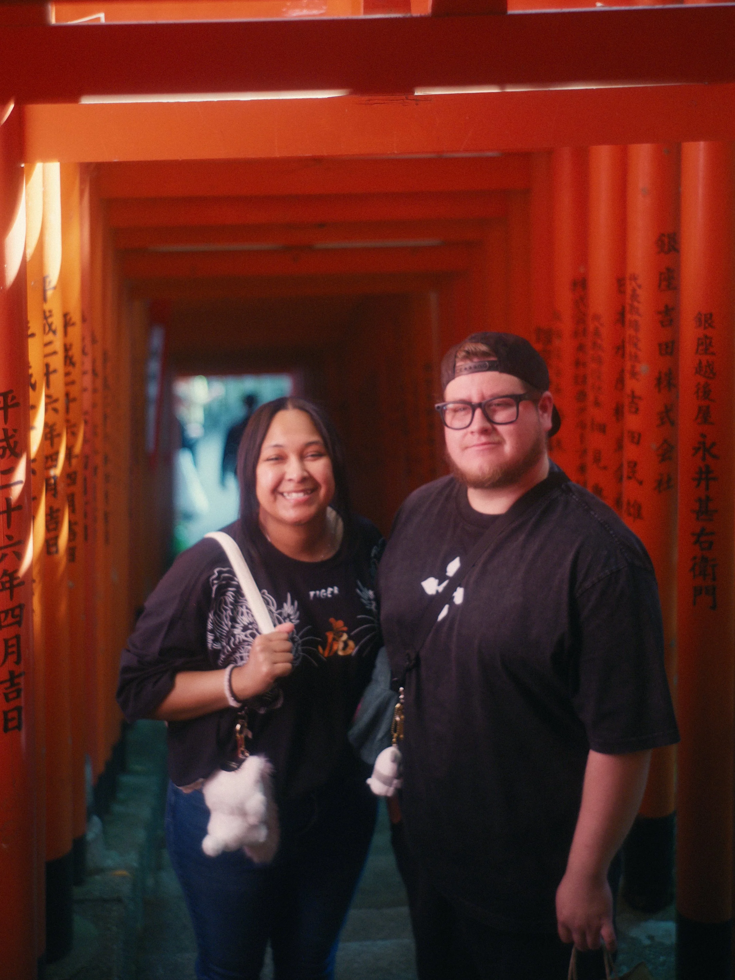 Two people standing under a series of orange torii gates at a Japanese shrine, smiling for the camera.