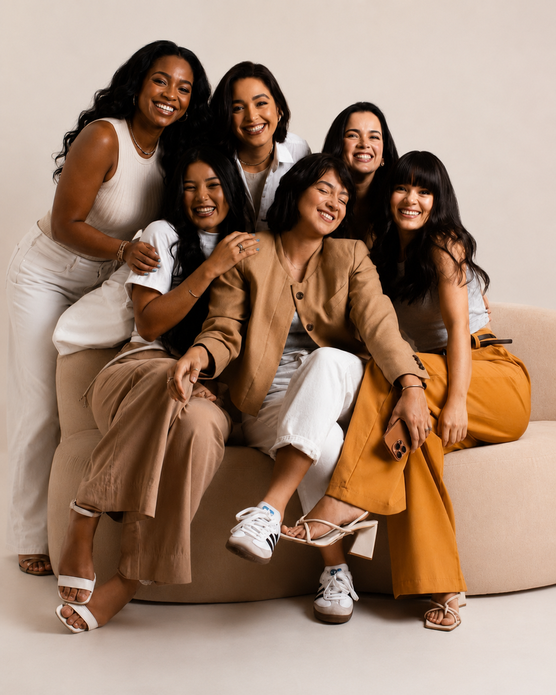 Group of six women smiling and sitting on a beige sofa against a plain background.