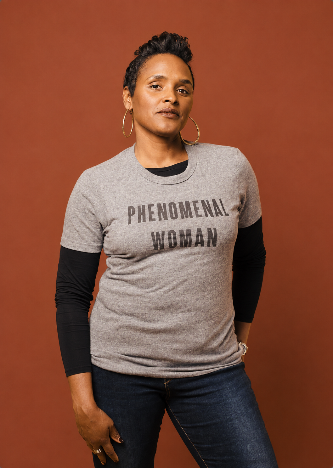 A confident woman with short curly hair wearing hoop earrings, a gray T-shirt that says 'PHENOMENAL WOMAN', a black long-sleeve shirt underneath, and dark jeans, standing against a brown background.