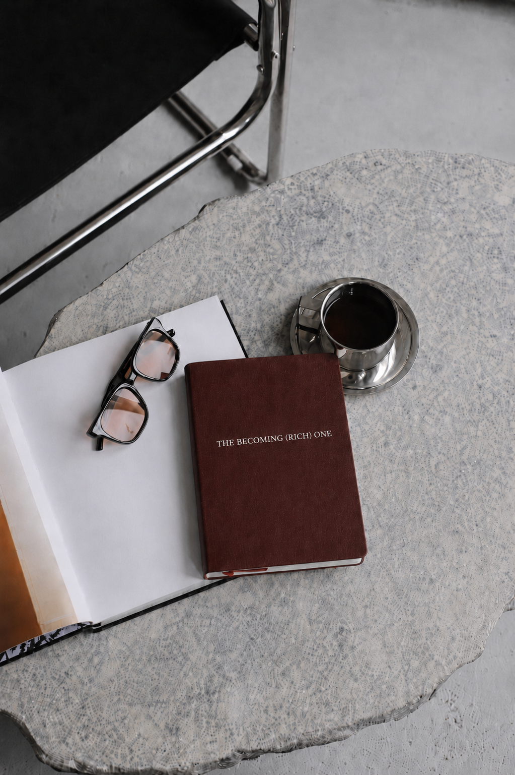 A table with a pair of glasses, an open book, a closed maroon book titled "The Becoming (Rich) One," a cup of dark coffee in a silver cup and saucer, all on a patterned white tablecloth. A chair is visible in the background.