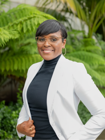 A woman with short black hair, glasses, and hoop earrings, wearing a black turtleneck and white blazer, standing outdoors with lush green tropical plants in the background.