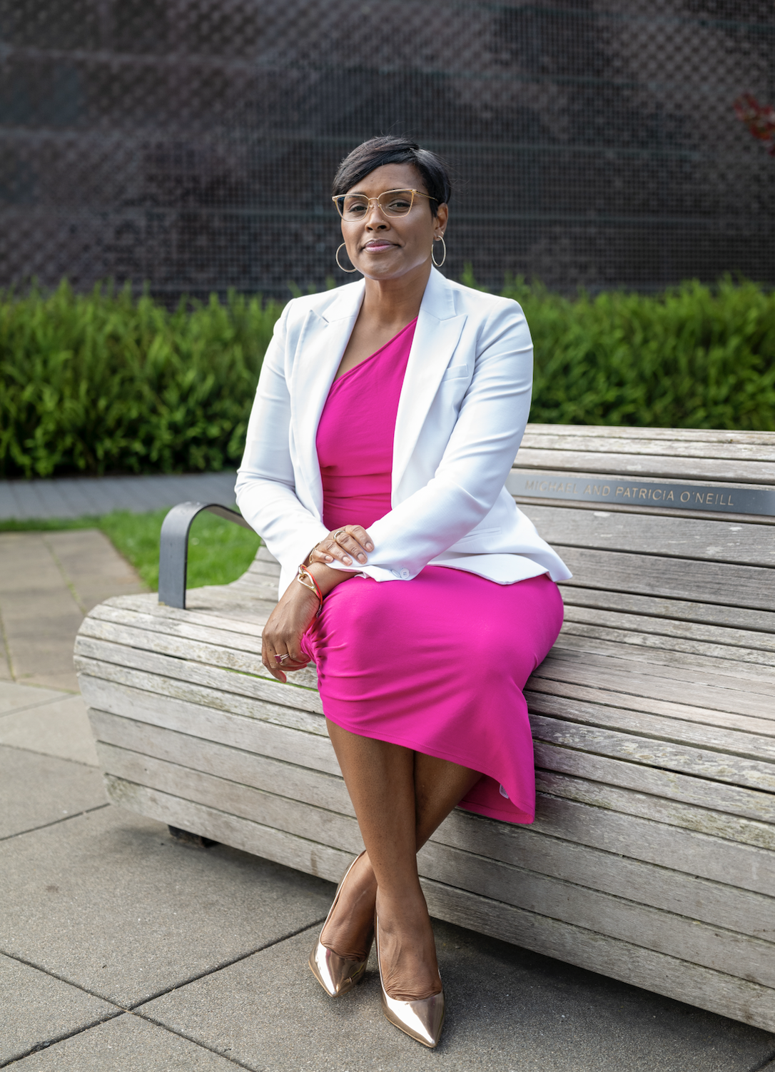 A woman sitting on a wooden park bench in an outdoor setting, wearing glasses, a white blazer, and a bright pink dress, with short black hair and hoop earrings.