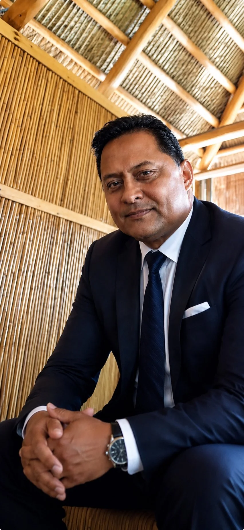 A man in a black suit and tie sitting indoors with a bamboo wall and roof structure in the background.
