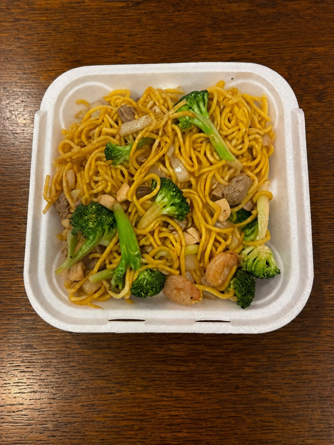 Container of stir-fried noodles with broccoli, chicken, and onions on a wooden table.
