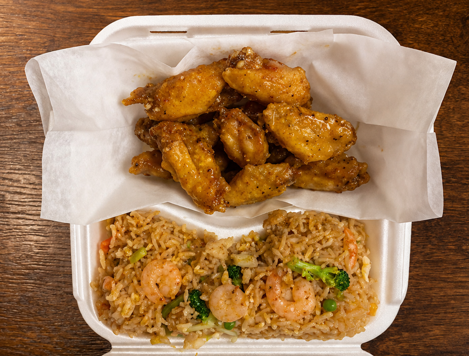 Fried chicken wings with sauce, served with fried rice containing shrimp, vegetables, and broccoli, in a takeout container on a wooden surface.