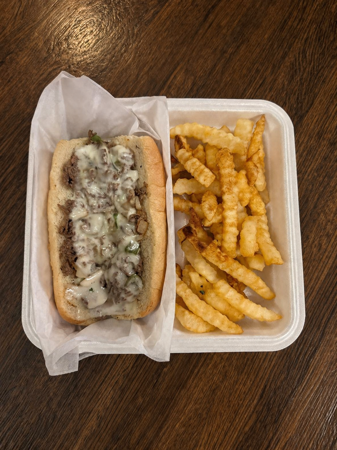 Hot dog with cheese, onions, and peppers in a bun, served with crinkle-cut French fries in a foam container on a wooden table.