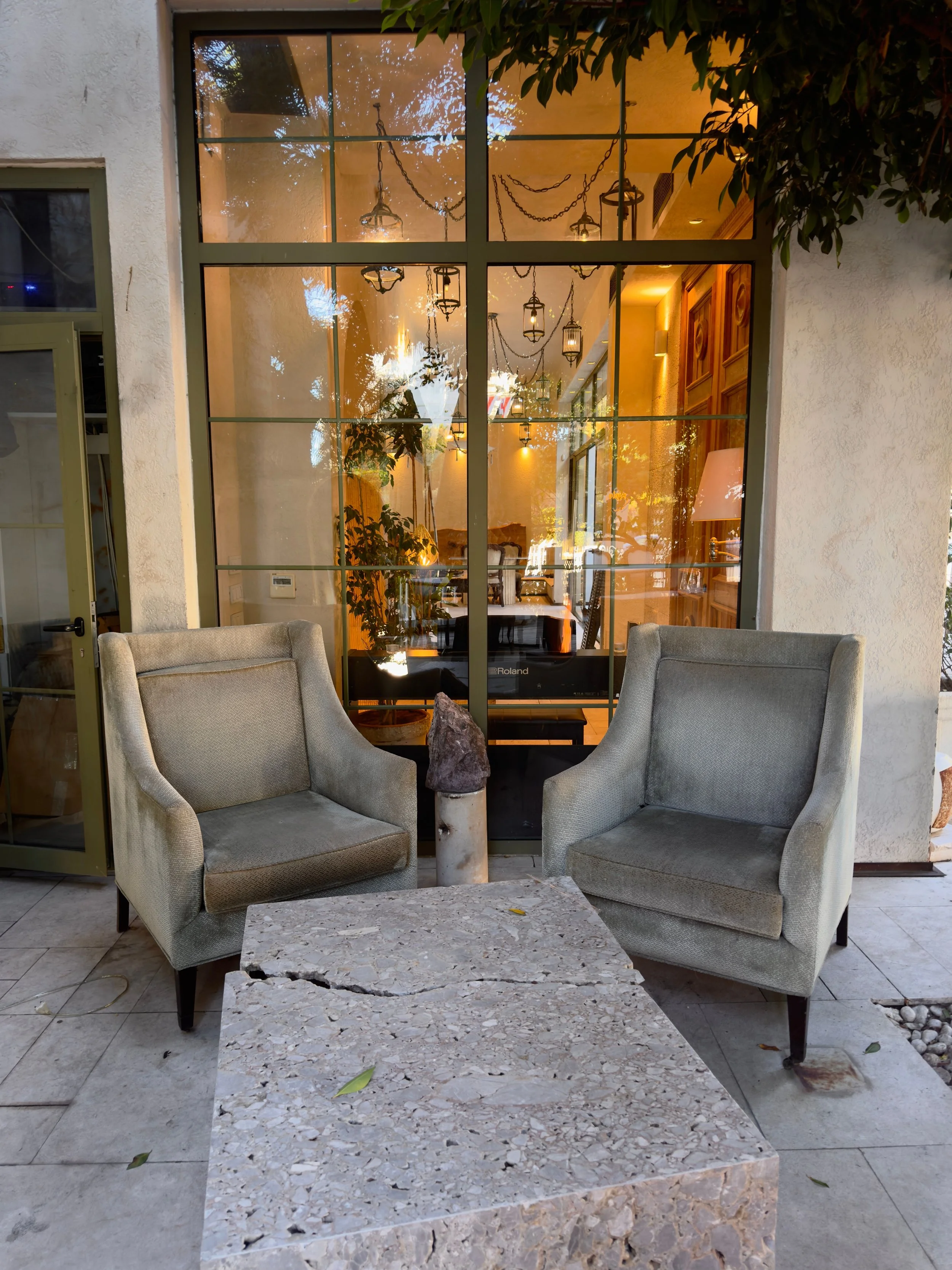 Two beige upholstered chairs surrounding a rectangular marble table with a rough natural edge, positioned outside in front of a large window with a view of a warmly lit interior of a dining area with hanging lanterns and chandeliers.