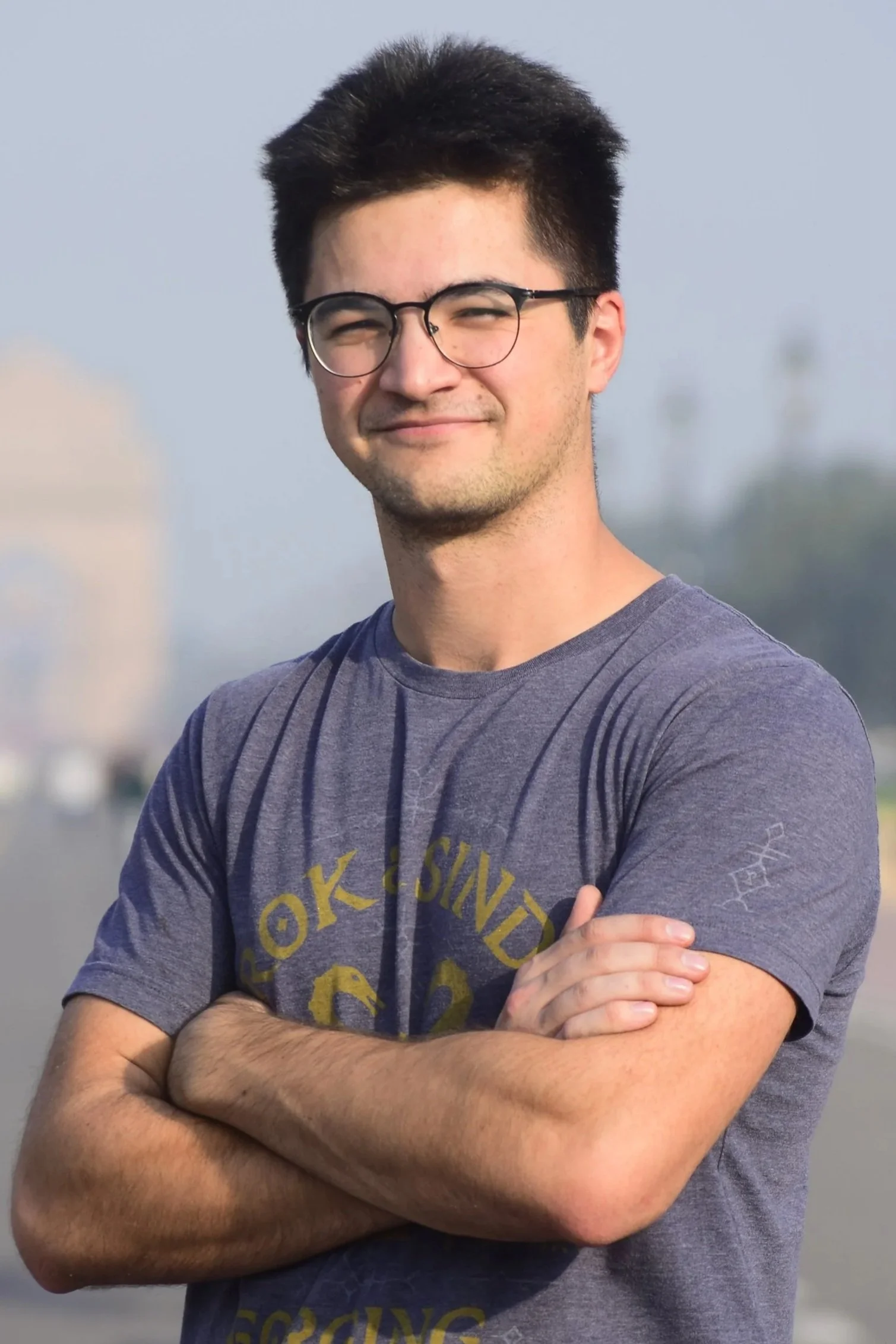 Young man with dark hair, glasses, wearing a gray t-shirt, arms crossed, outdoors with a blurred background.