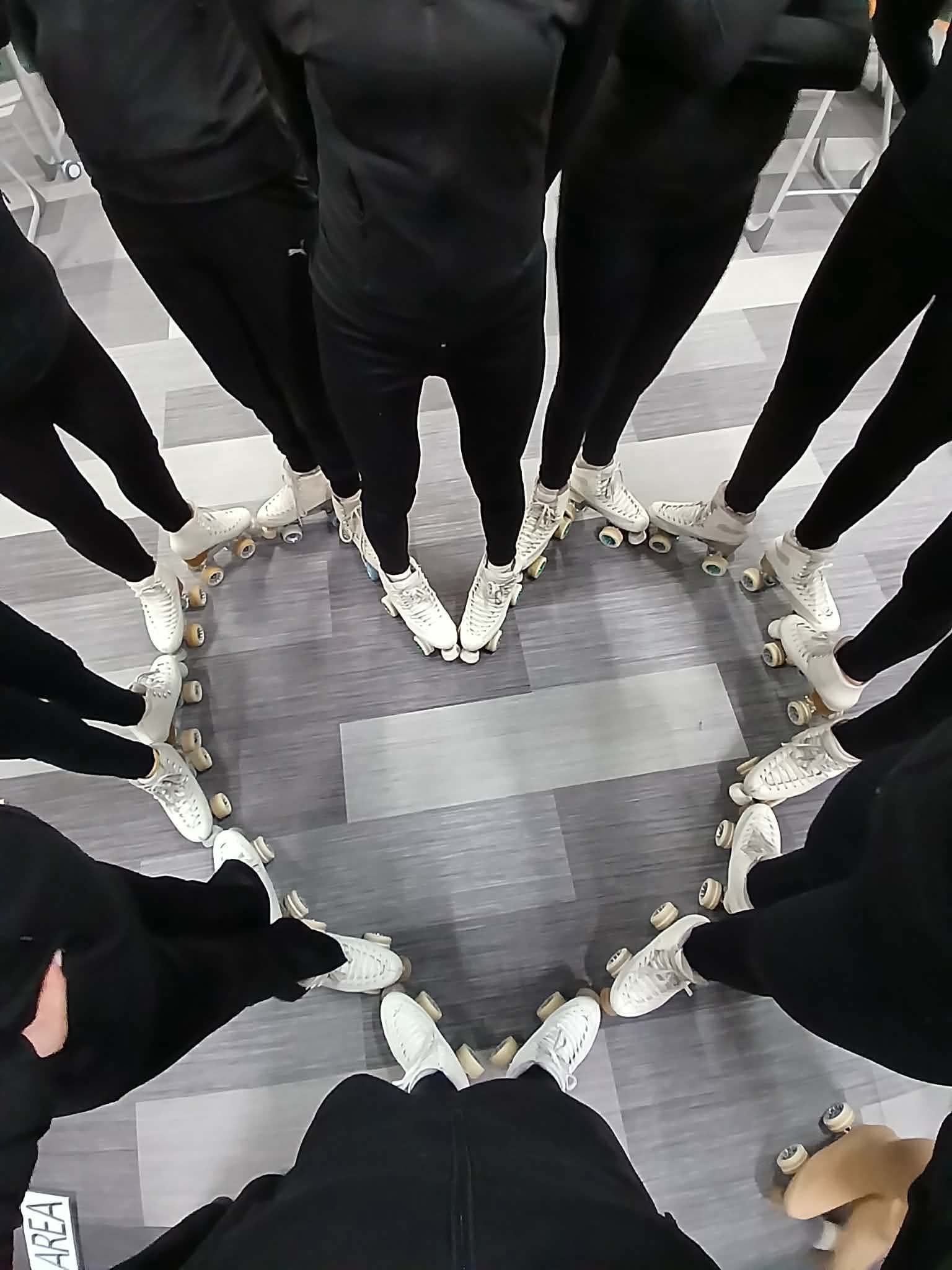 A group of people on roller skates standing in a circle, viewed from above, with their ankles touching and wearing black clothing.
