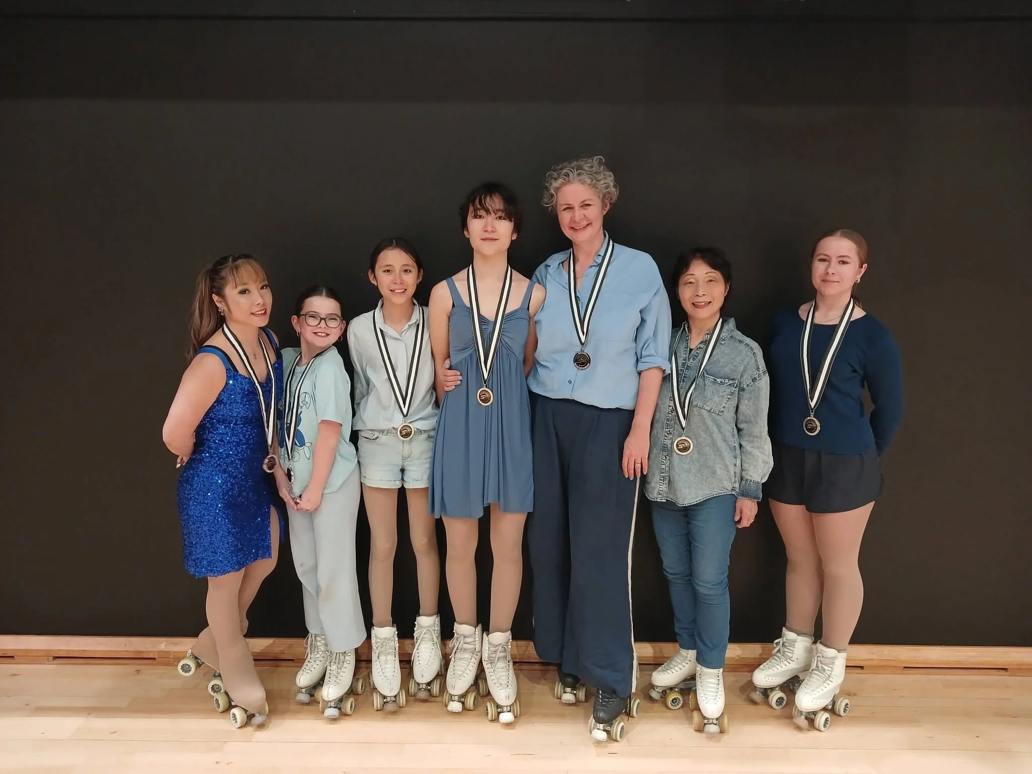 Group of club members in roller skates, wearing medals, standing against a dark wall, smiling for a photo after a roller skating event.