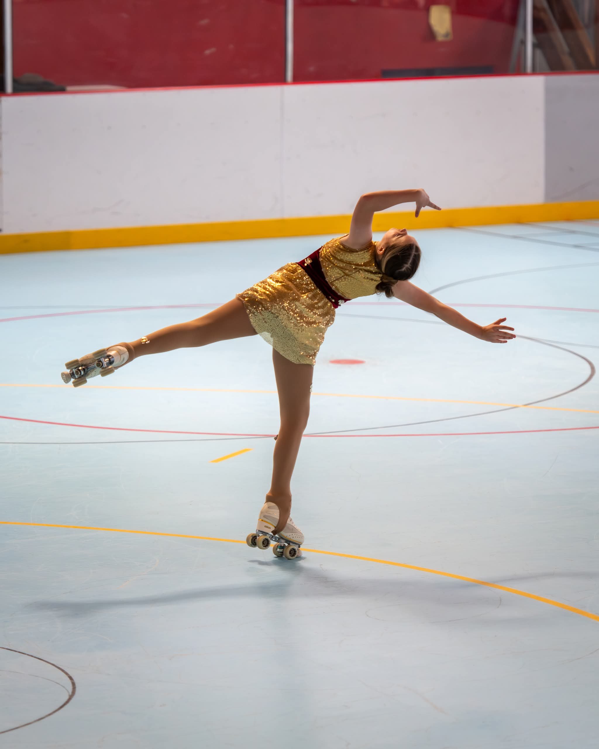A young girl roller skating on an indoor rink, wearing a shiny gold dress, with her arm extended and one leg lifted in a graceful pose.