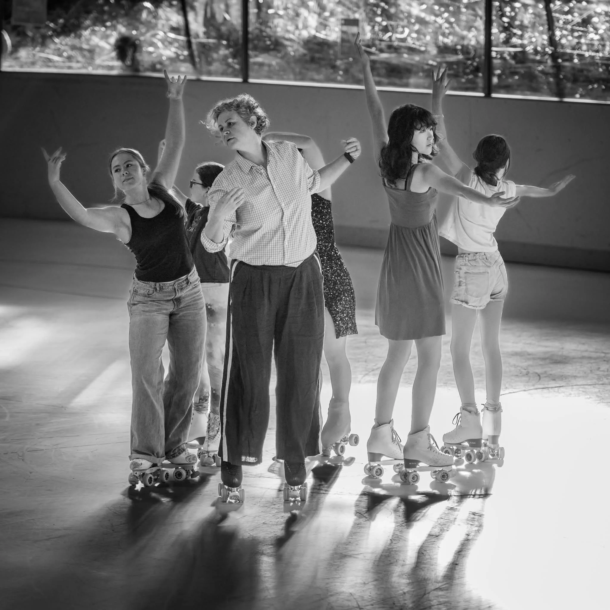 A group of people on roller skates, skating indoors near large windows, with sunlight filtering through outside windows.