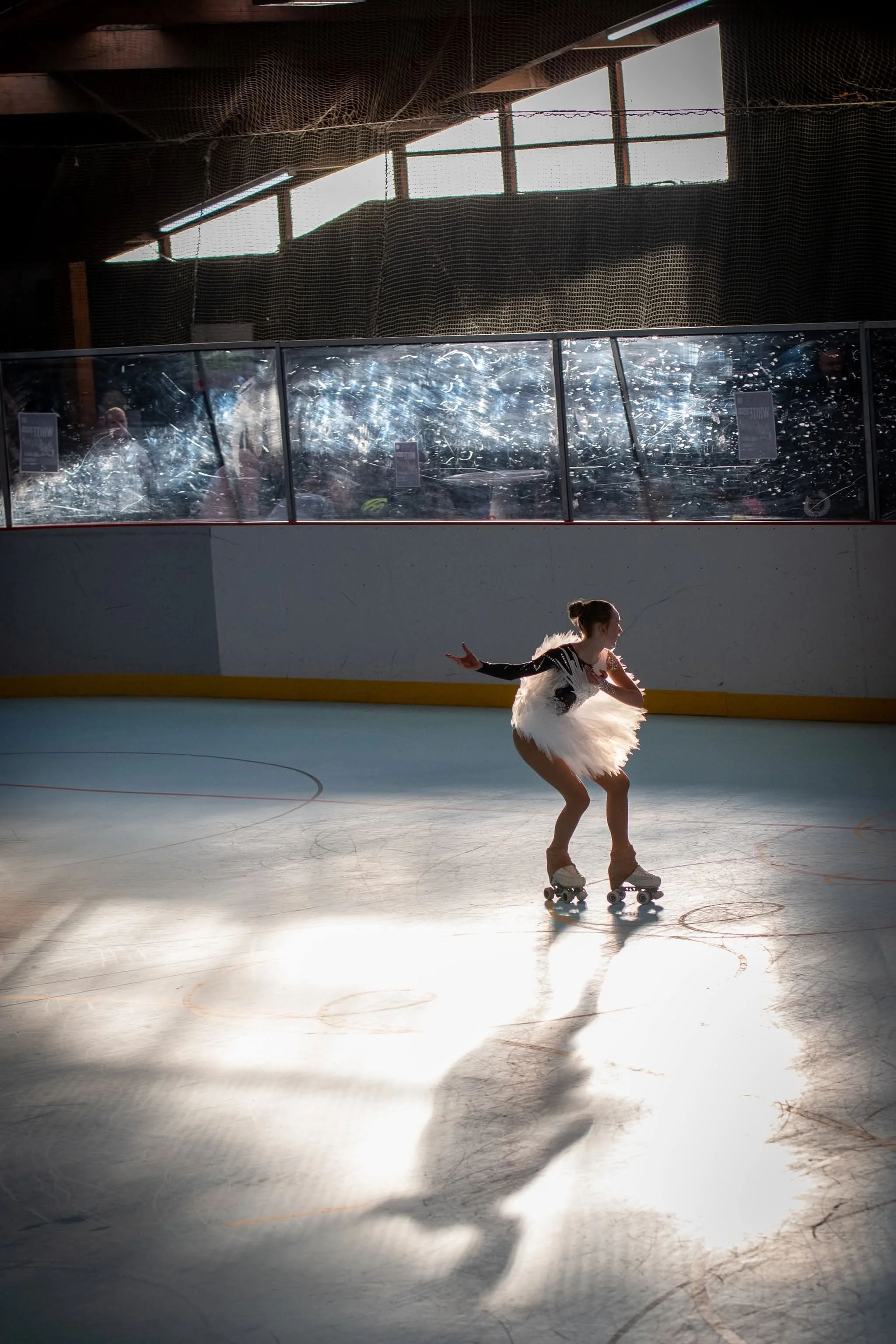 A young girl in a white figure skating costume is performing on an indoor skating rink, illuminated by sunlight coming through windows above. She is on roller skates, skating in a pose with arms extended, and her shadow is visible on the floor.