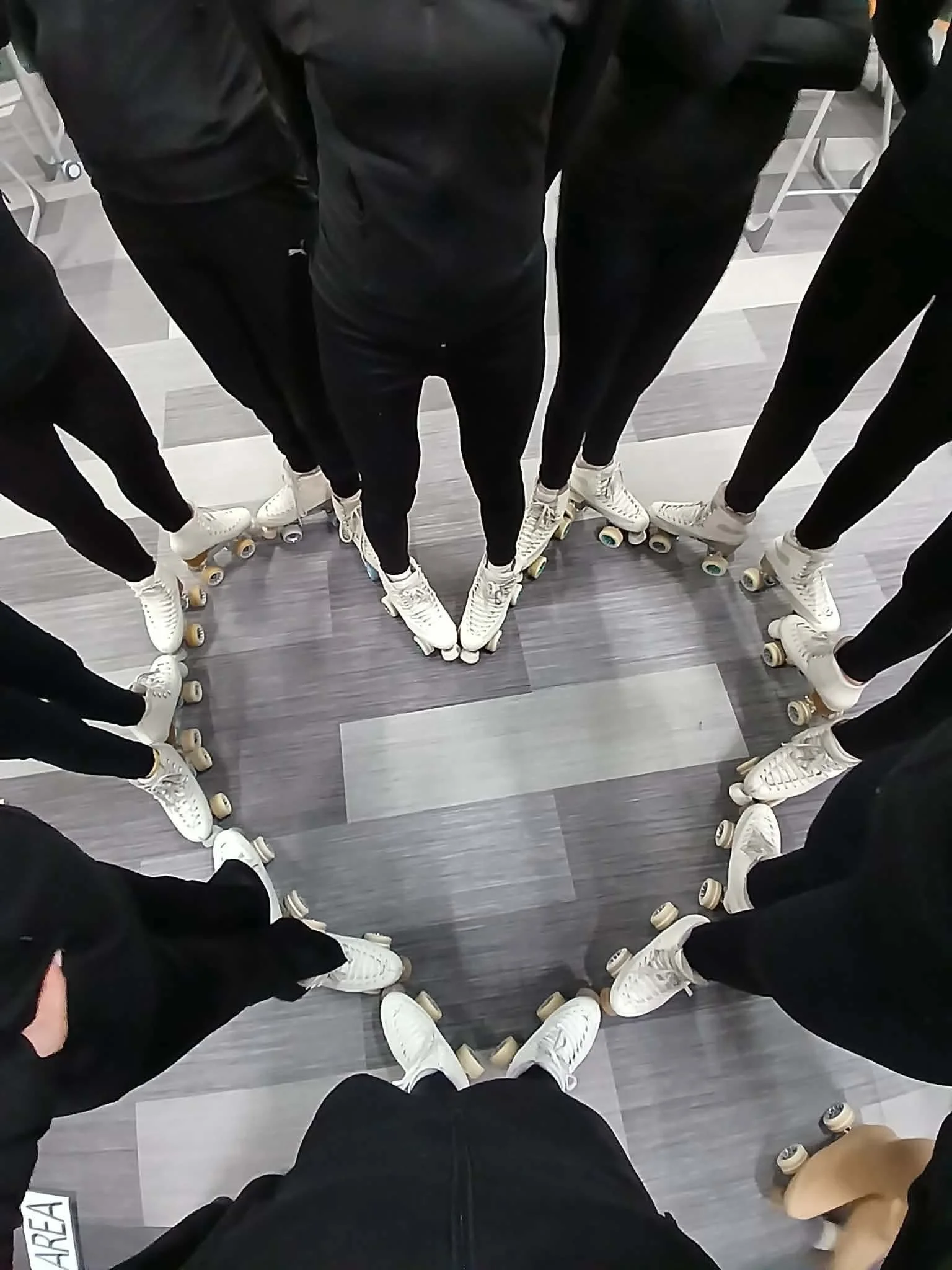 People standing in a circle on roller skates, viewed from below, with gray flooring and chairs in the background.