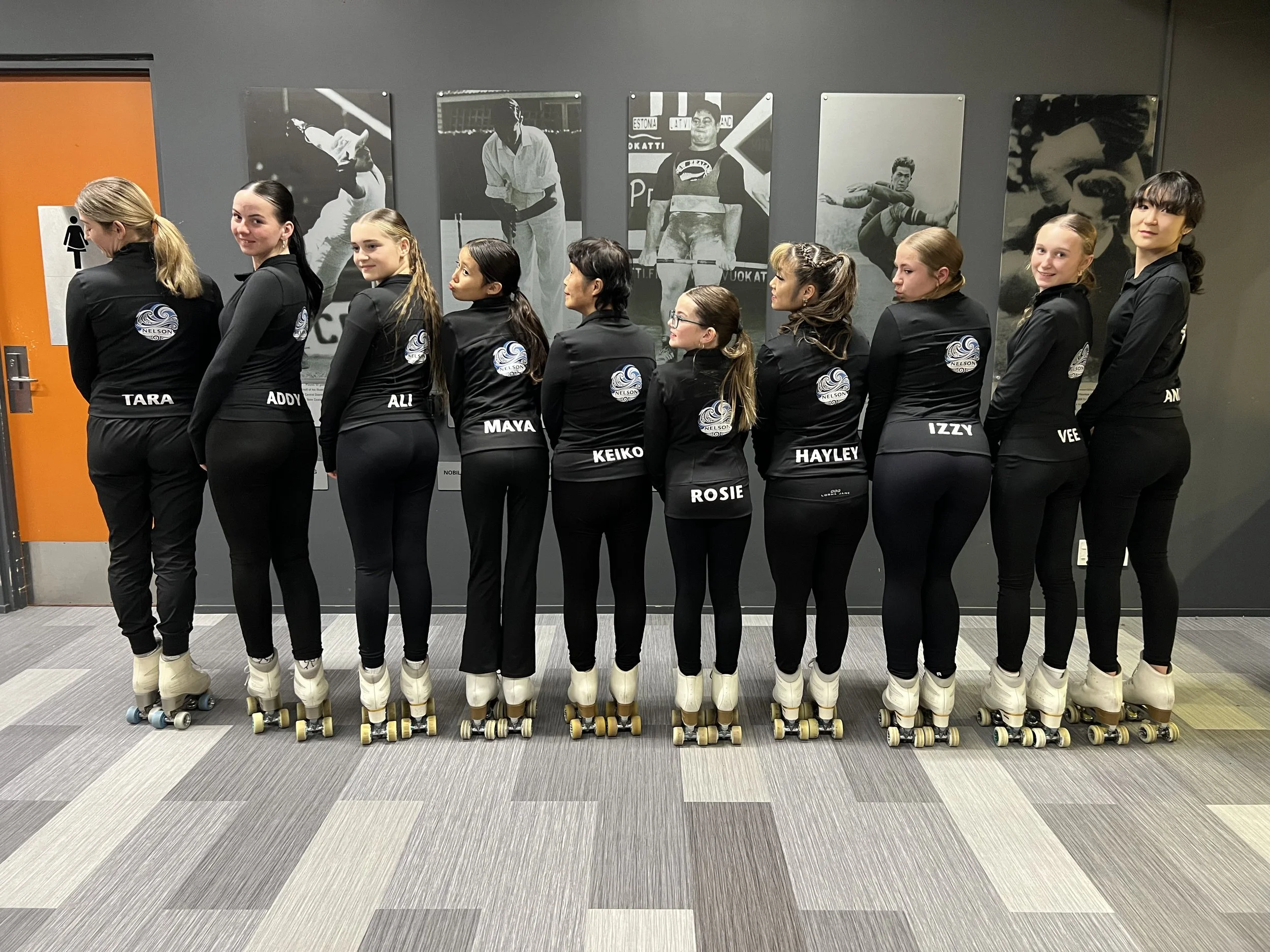 A group of nine young women wearing black jackets with a logo on the back, black pants, and roller skates, standing in a line facing away from the camera in an indoor skating rink. They have different hairstyles and are positioned in front of a wall with black-and-white sports posters.