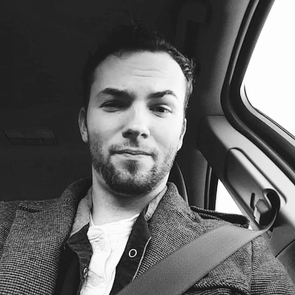 Black and white photo of a man sitting in a vehicle, wearing a seatbelt, with a window to his right showing bright sky outside.