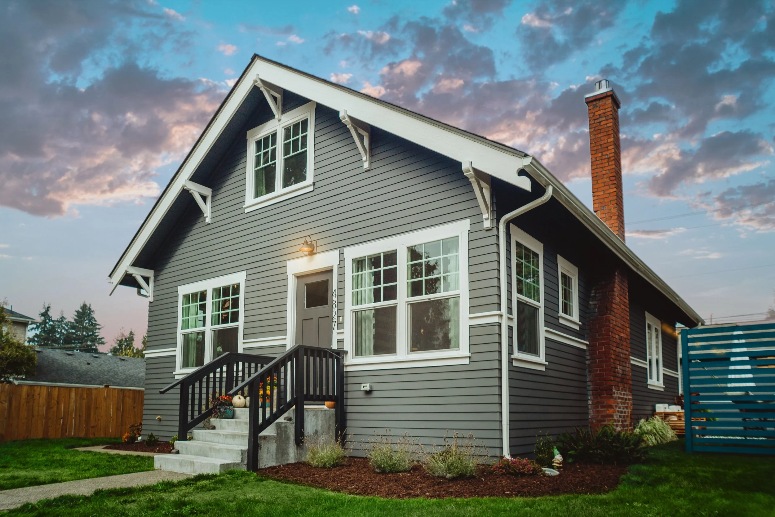 A two-story house with gray siding, white trim, a black front door, and large windows. The house has a front porch with stairs, black railings, and outdoor decorations including pumpkins. There is a brick chimney on the right side and a wooden fence on the left. The sky is partly cloudy with shades of blue and pink, indicating sunset or sunrise.