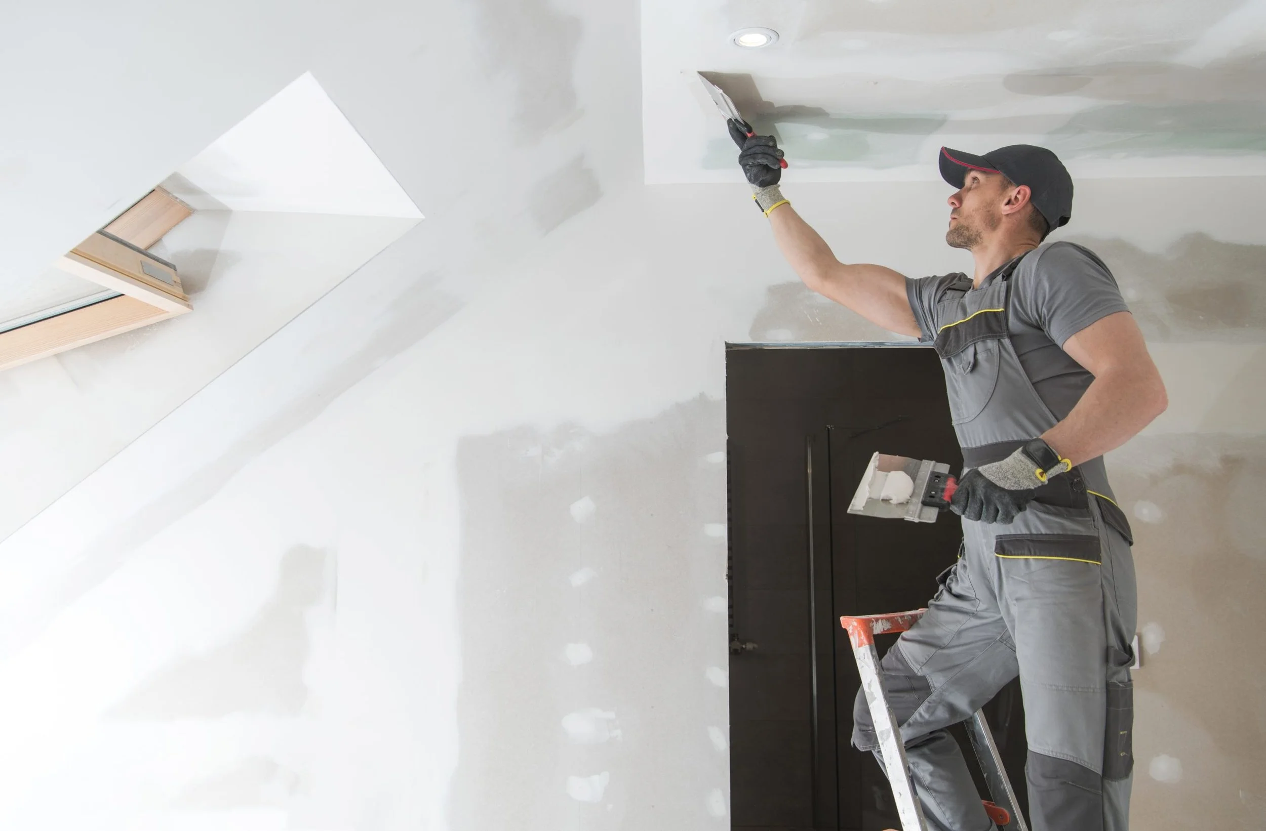 A man in work overalls and a cap standing on a ladder, smoothing out drywall on a ceiling with a trowel.
