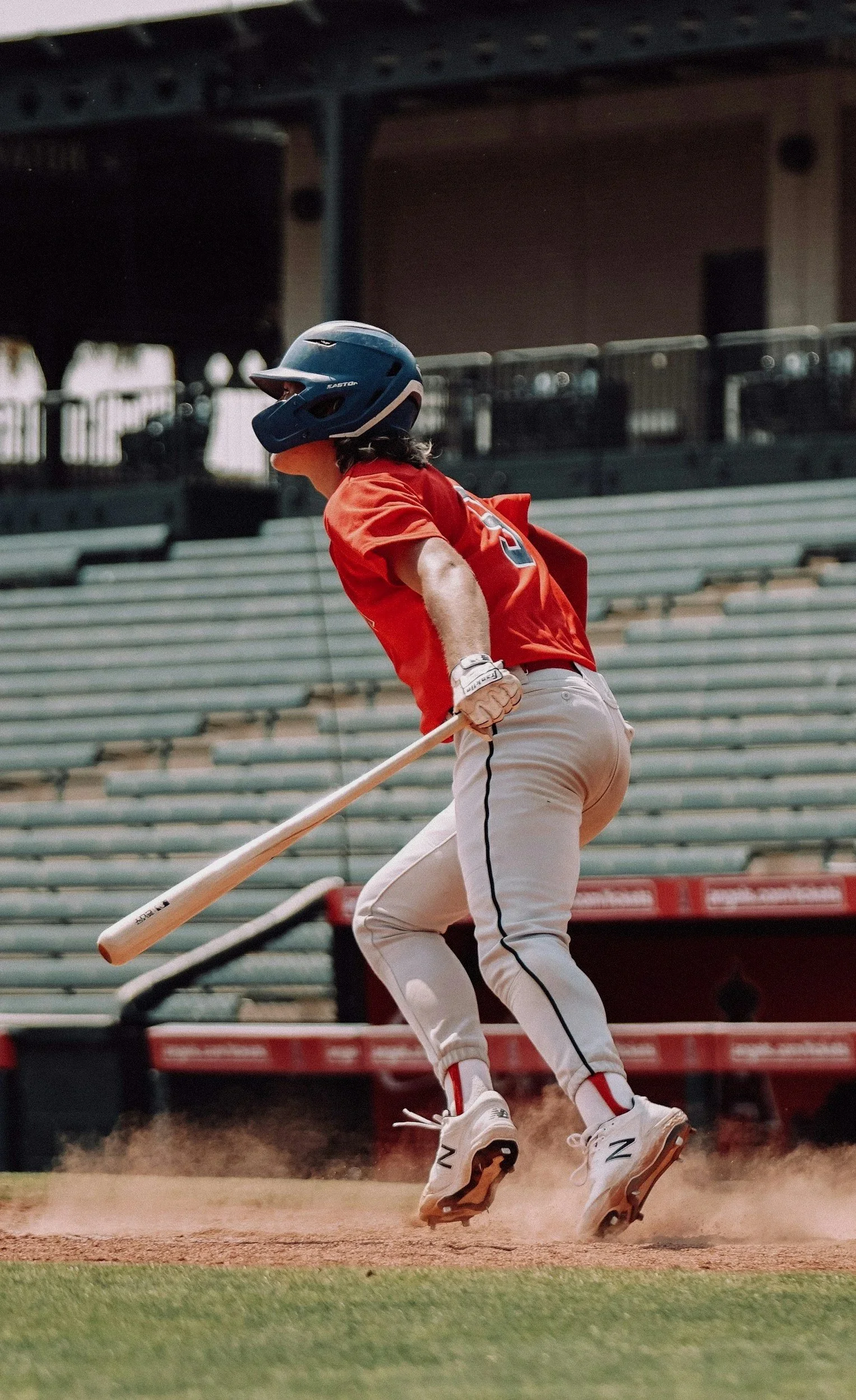 A baseball player in a red jersey and white pants running or sliding on the field with a baseball bat in hand, wearing a blue helmet, with empty stadium seating in the background.