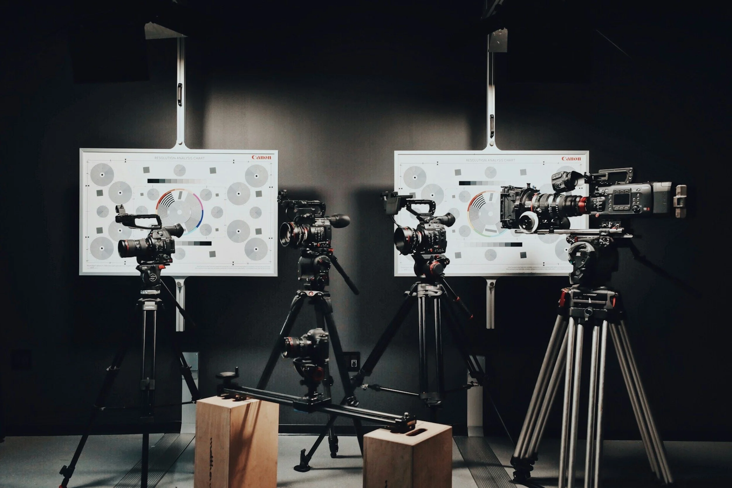 Three DSLR cameras on tripods facing white charts with gray circles and color bars, set against a black background in a photography studio.