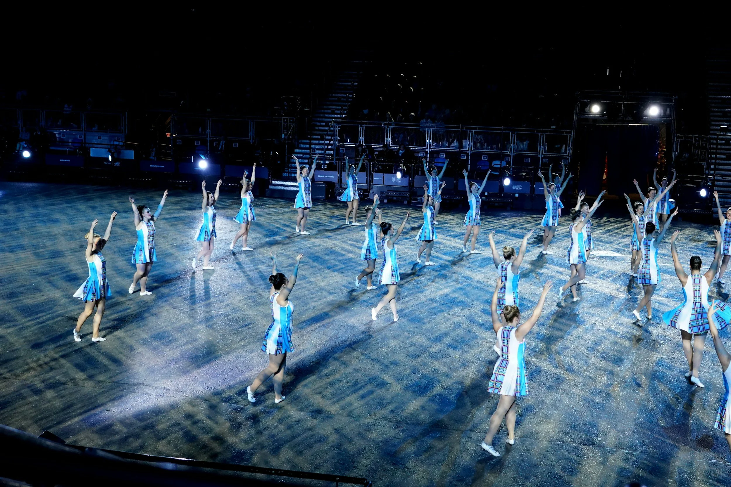 Group of synchronized dancers in plaid dresses performing on a stage in front of an audience, with spotlights shining on them.
