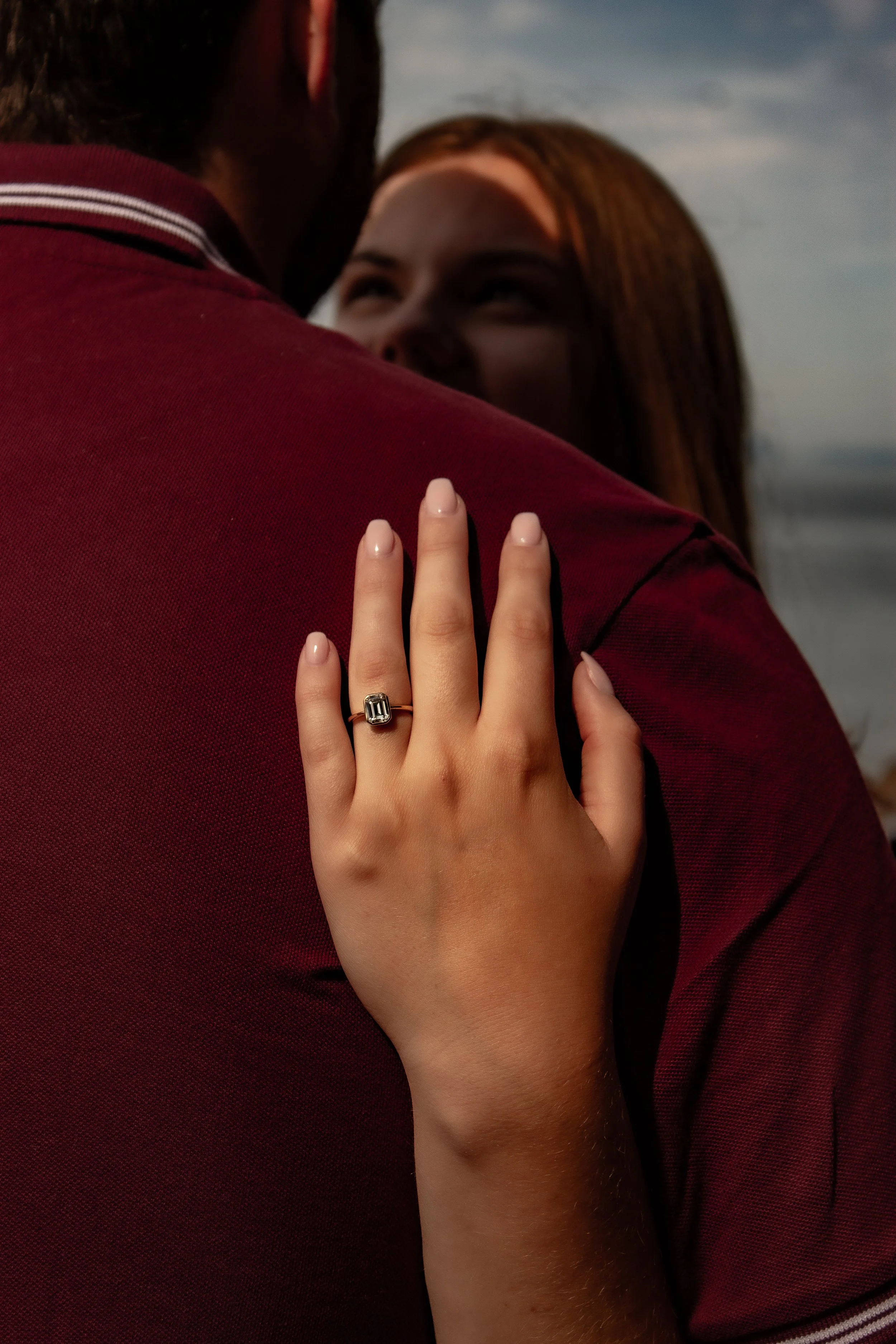 A woman with light skin and red hair smiling as she embraces a man in a maroon shirt at the beach during sunset.