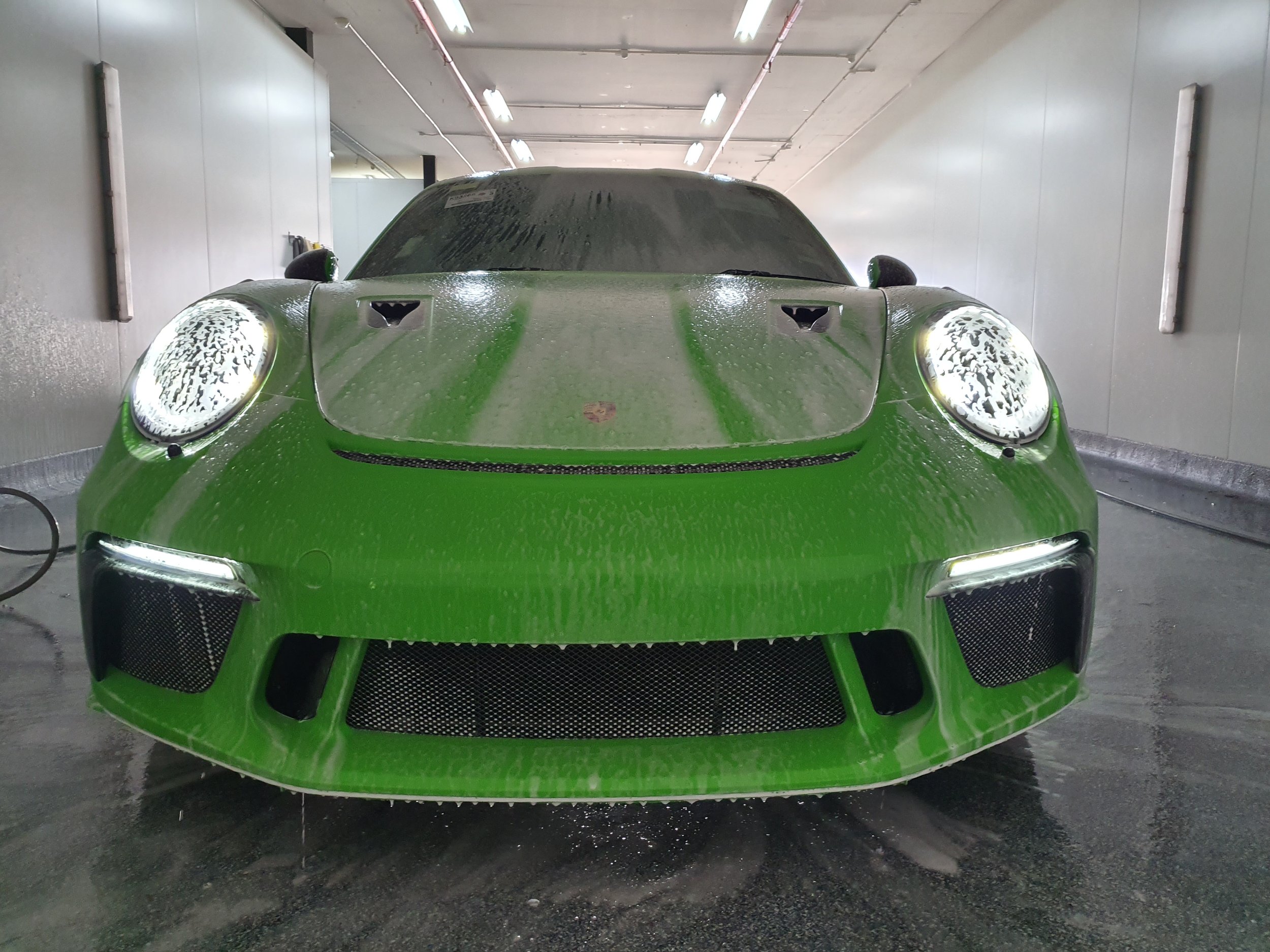 A bright green sports car with wet surface in an indoor car wash area, with high ceilings, fluorescent lighting, and water droplets on the car's surface.