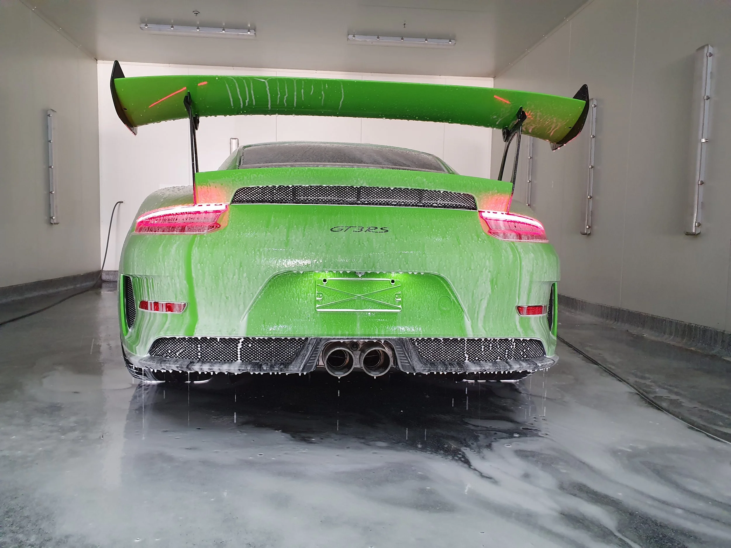 Green Porsche GT3 RS sports car in a car wash or detailing center, with water and soap visible on its surface, and a large rear spoiler.