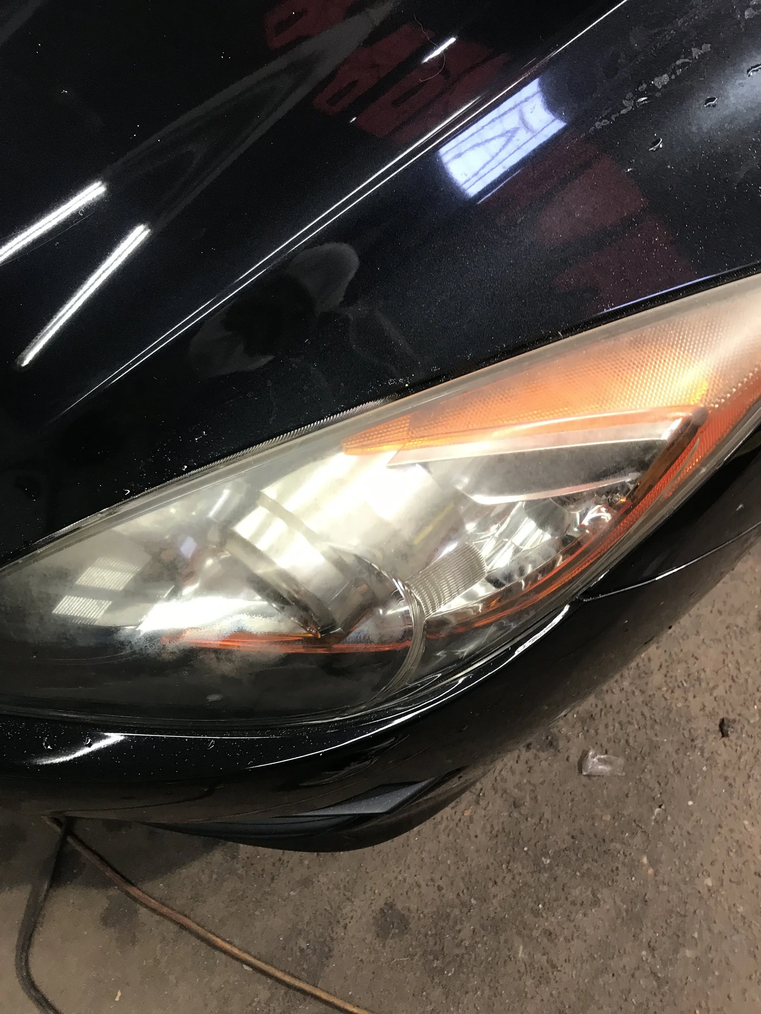 Close-up of the front of a black car, showing the headlight and part of the hood, with some water droplets on the surface, parked on a dirt or gravel surface.