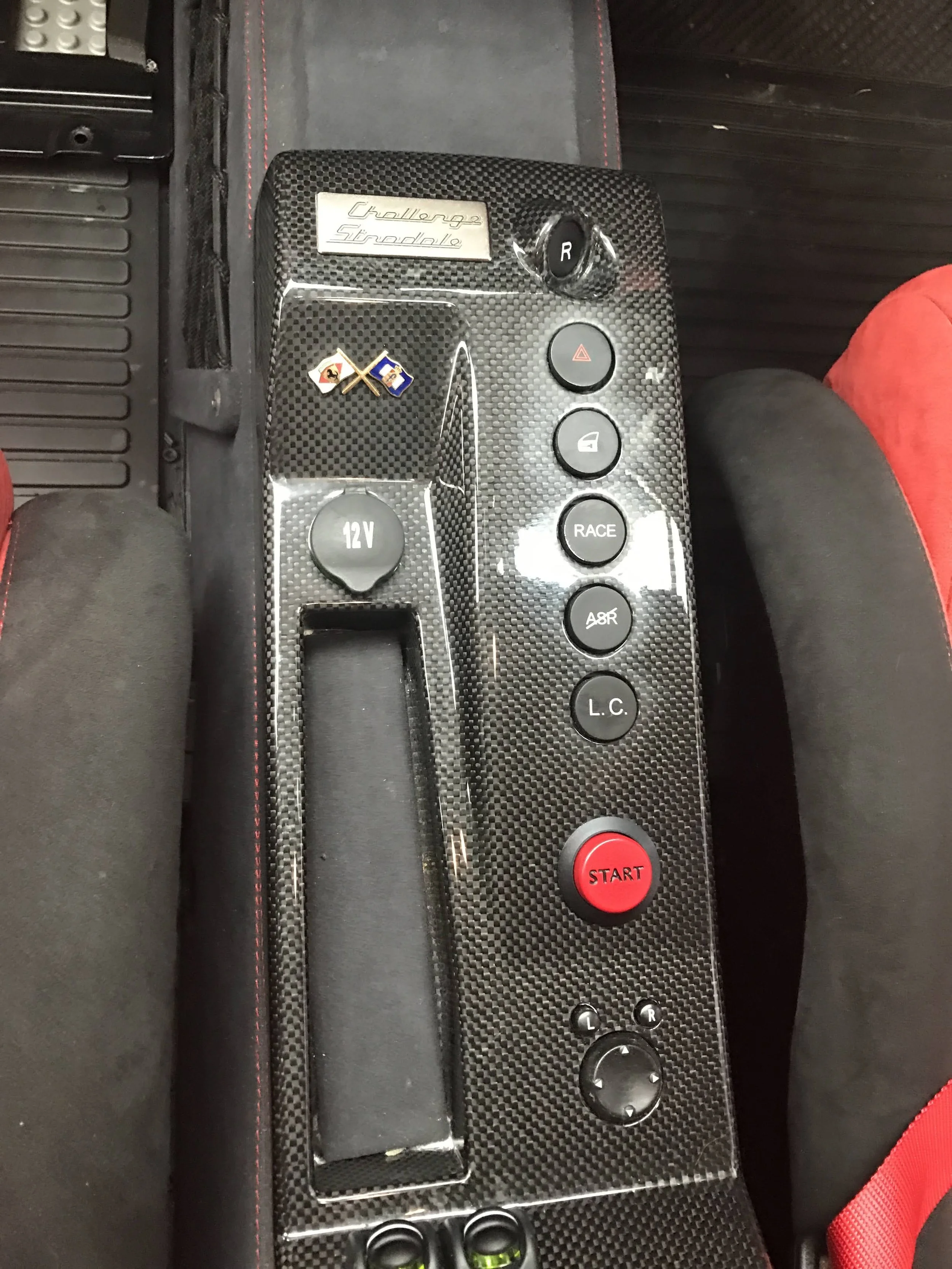 Close-up of a carbon fiber panel in a sports car interior with buttons labeled R, hazard, door lock, RACE, AER, L.C., L, R, and a large red START button. The panel has a euro race badge and a 12V power socket.