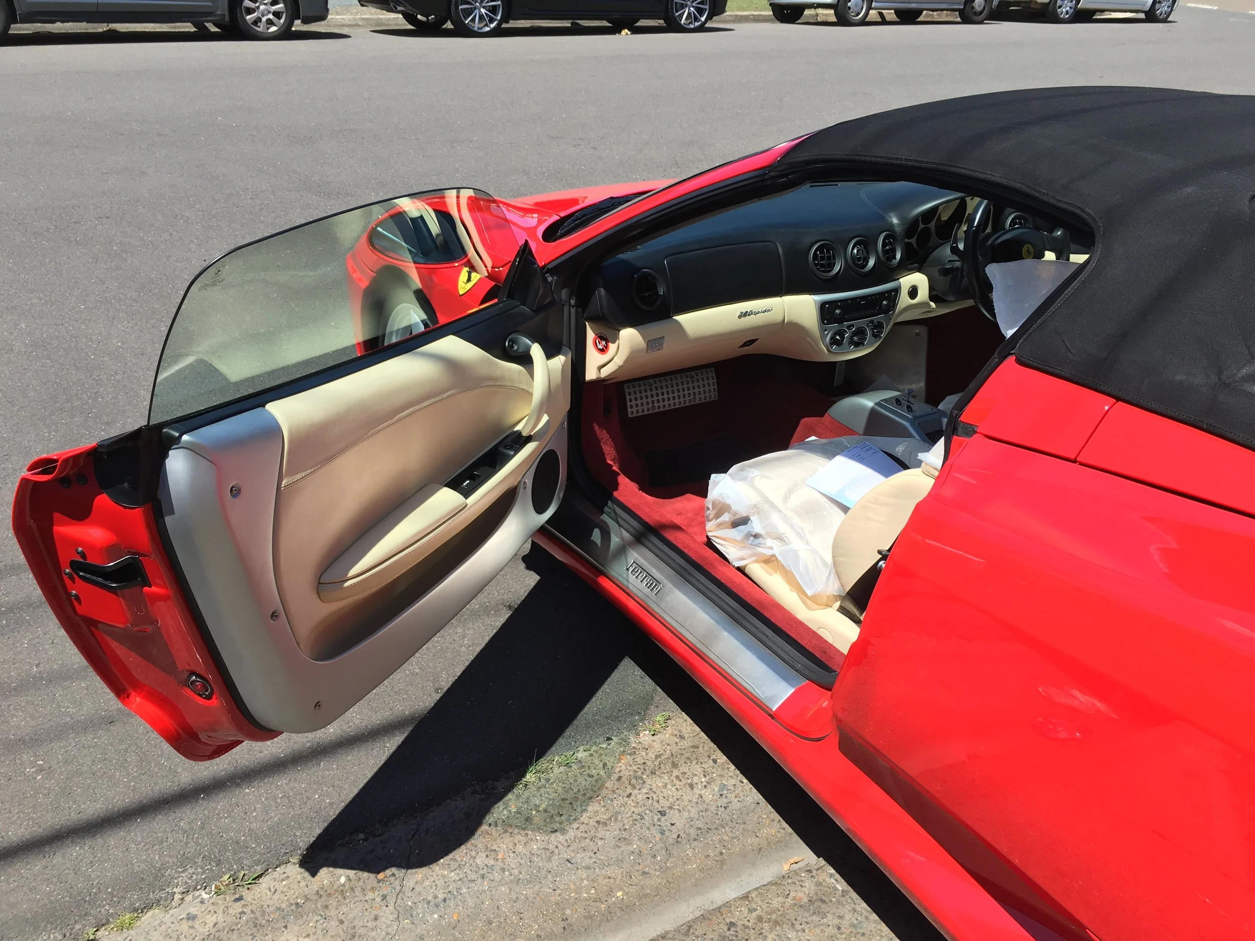 A red convertible sports car with a black soft top, parked in a parking lot with other vehicles in the background. The driver's side door is open, revealing a beige interior and part of the dashboard.