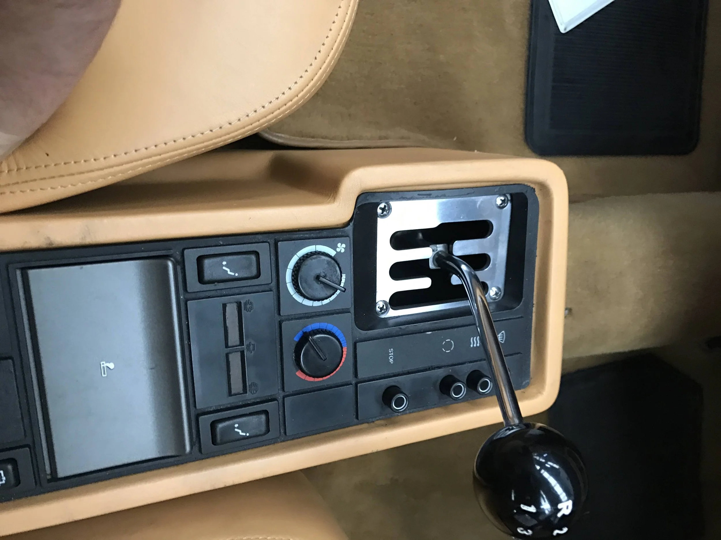 A close-up of a car's center console showing climate control dials, a gear shift lever, and a portion of a beige seat and brown carpet