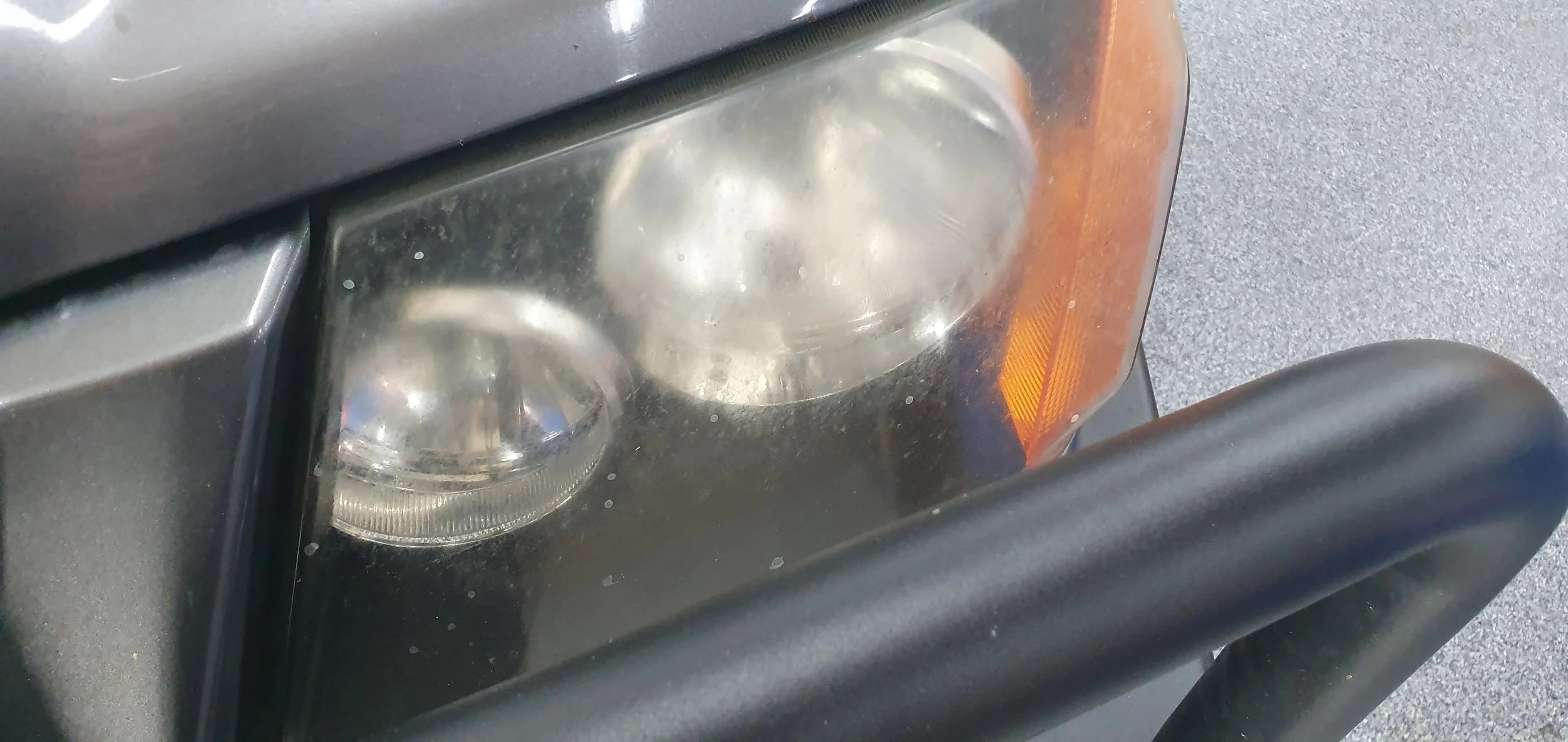Close-up of a vehicle's headlight with a foggy or cloudy appearance, surrounded by a black frame and an orange side marker light on the right. Part of the black bumper is visible at the bottom of the image.