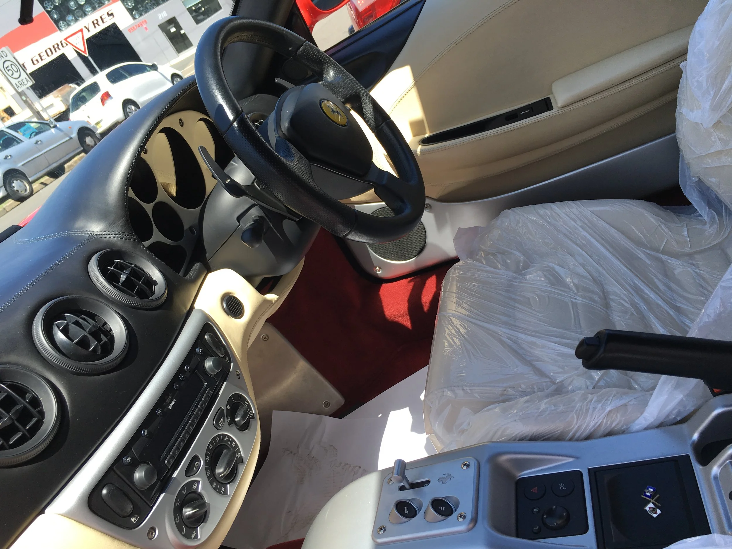 The interior of a luxury car showing the dashboard, steering wheel with a Ferrari logo, beige leather seats covered with plastic, and an outside view of parked cars and street signs.