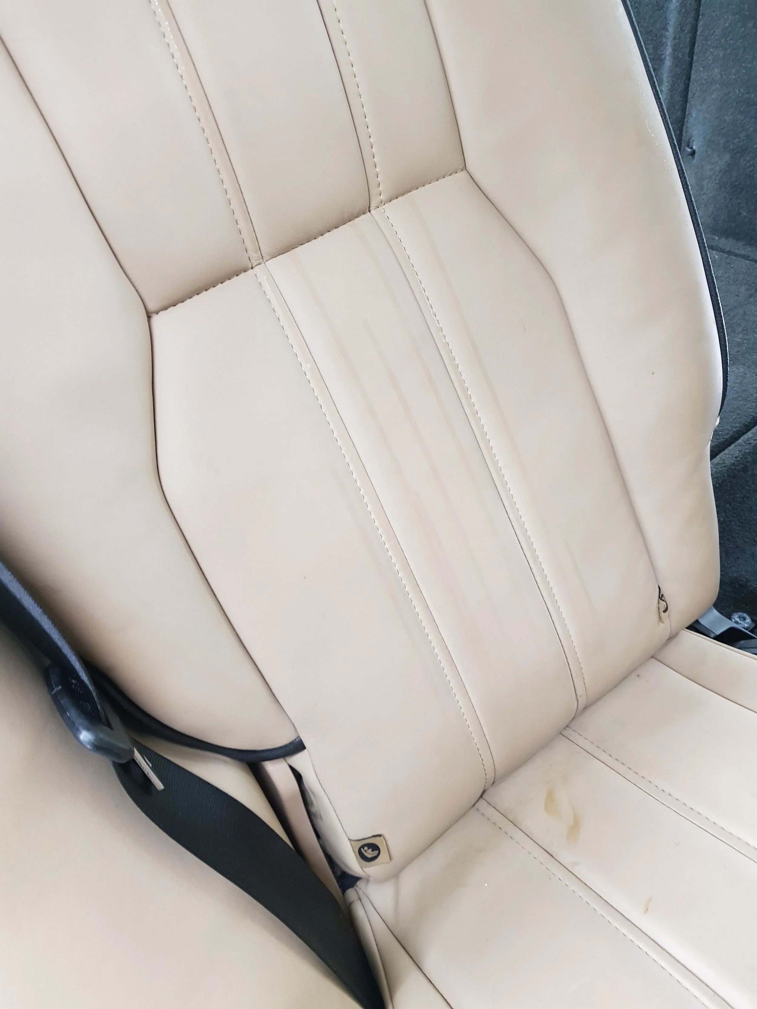 Close-up of beige leather car seat with visible stitching, near a beige interior wall with some stains.