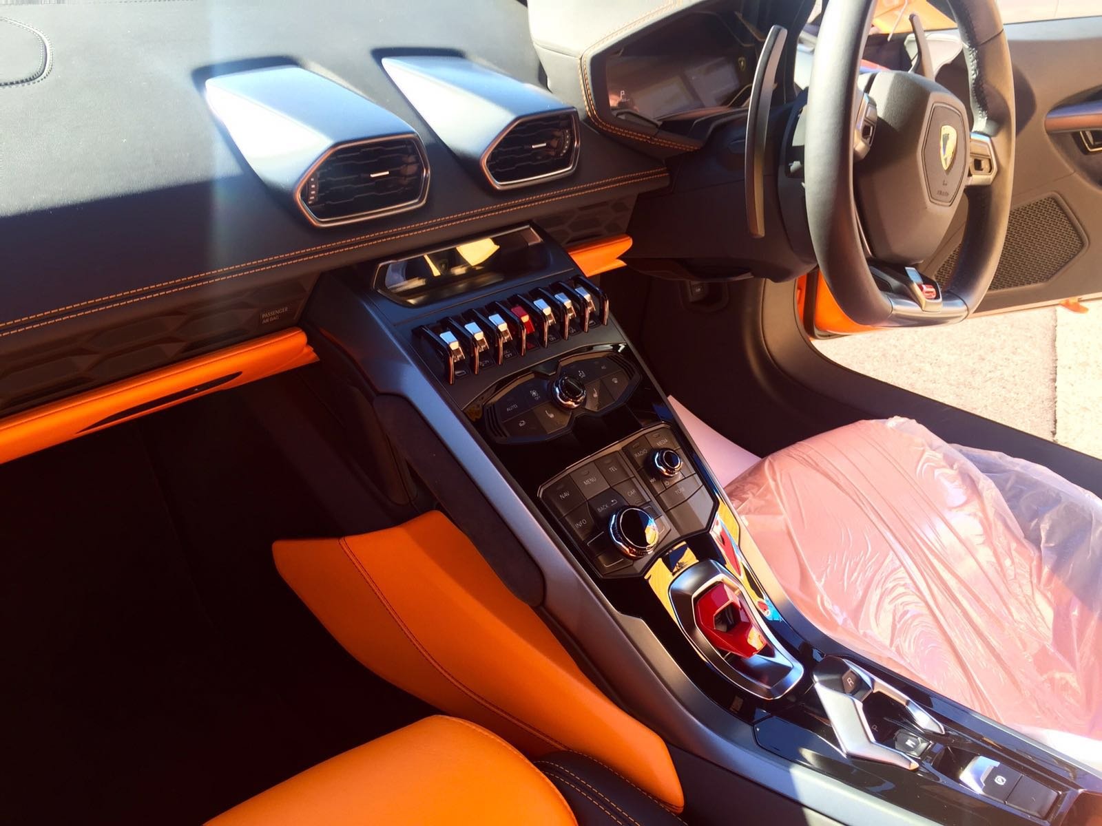 Interior of a luxury sports car, showing the dashboard, central control area with gear shifter, and orange leather seats, with a plastic cover on the passenger seat.