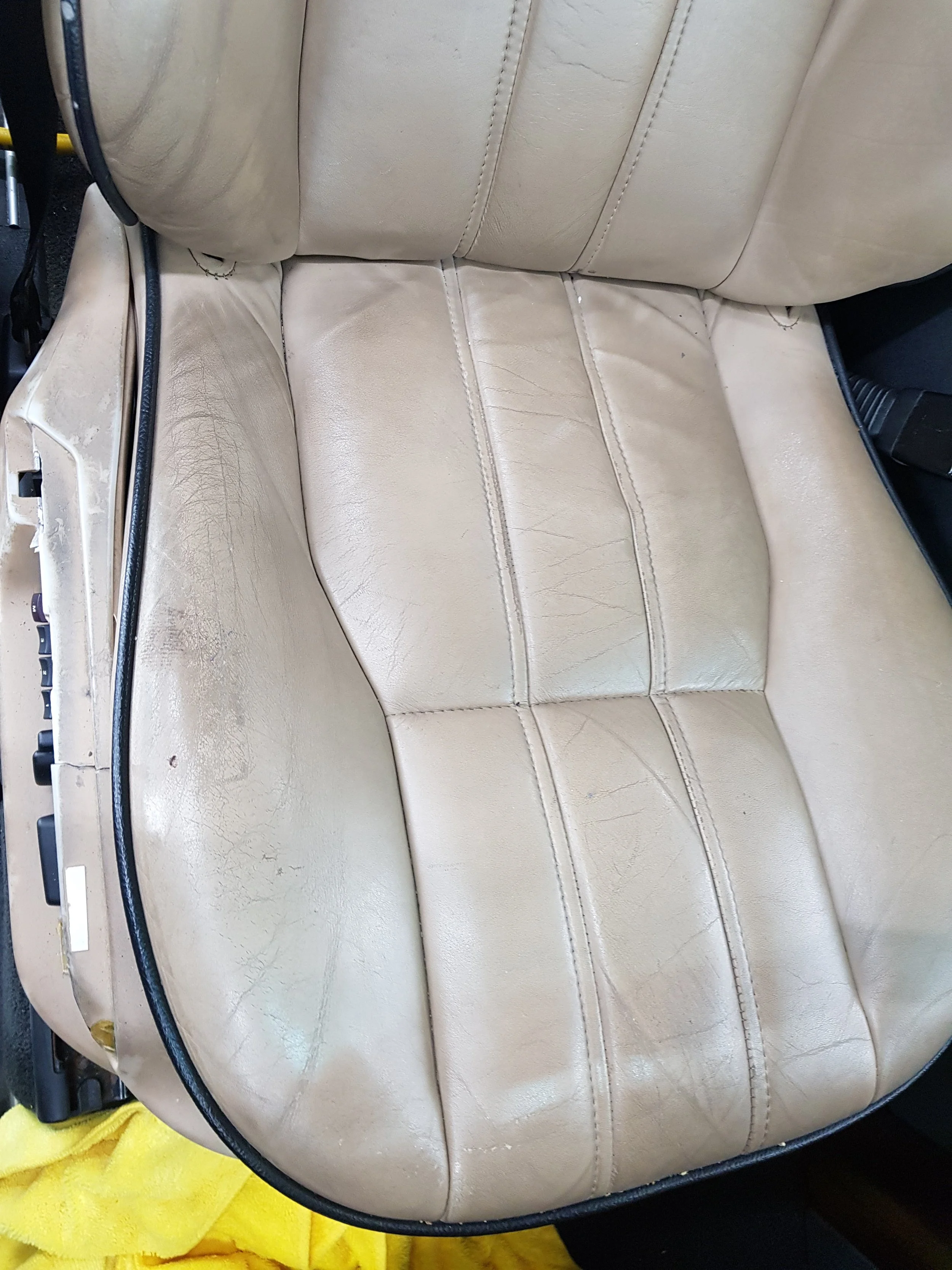 Close-up of a worn, beige leather car seat with visible cracks and creases, and a yellow cloth underneath.