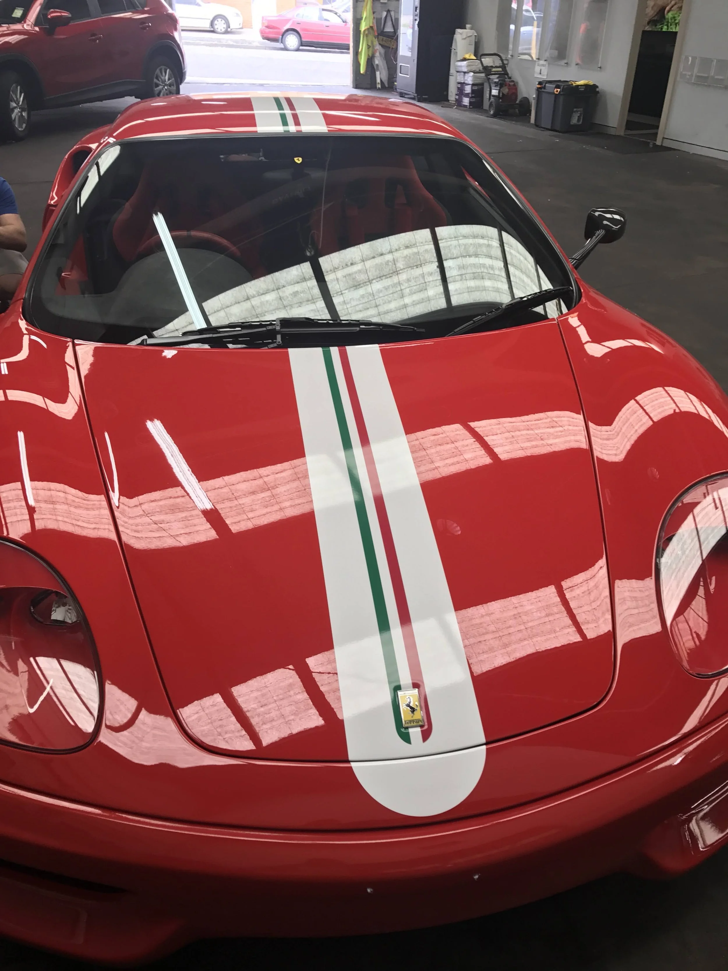 Red sports car with a white and green racing stripe, parked indoors, with a Ferrari logo emblem visible at the front.