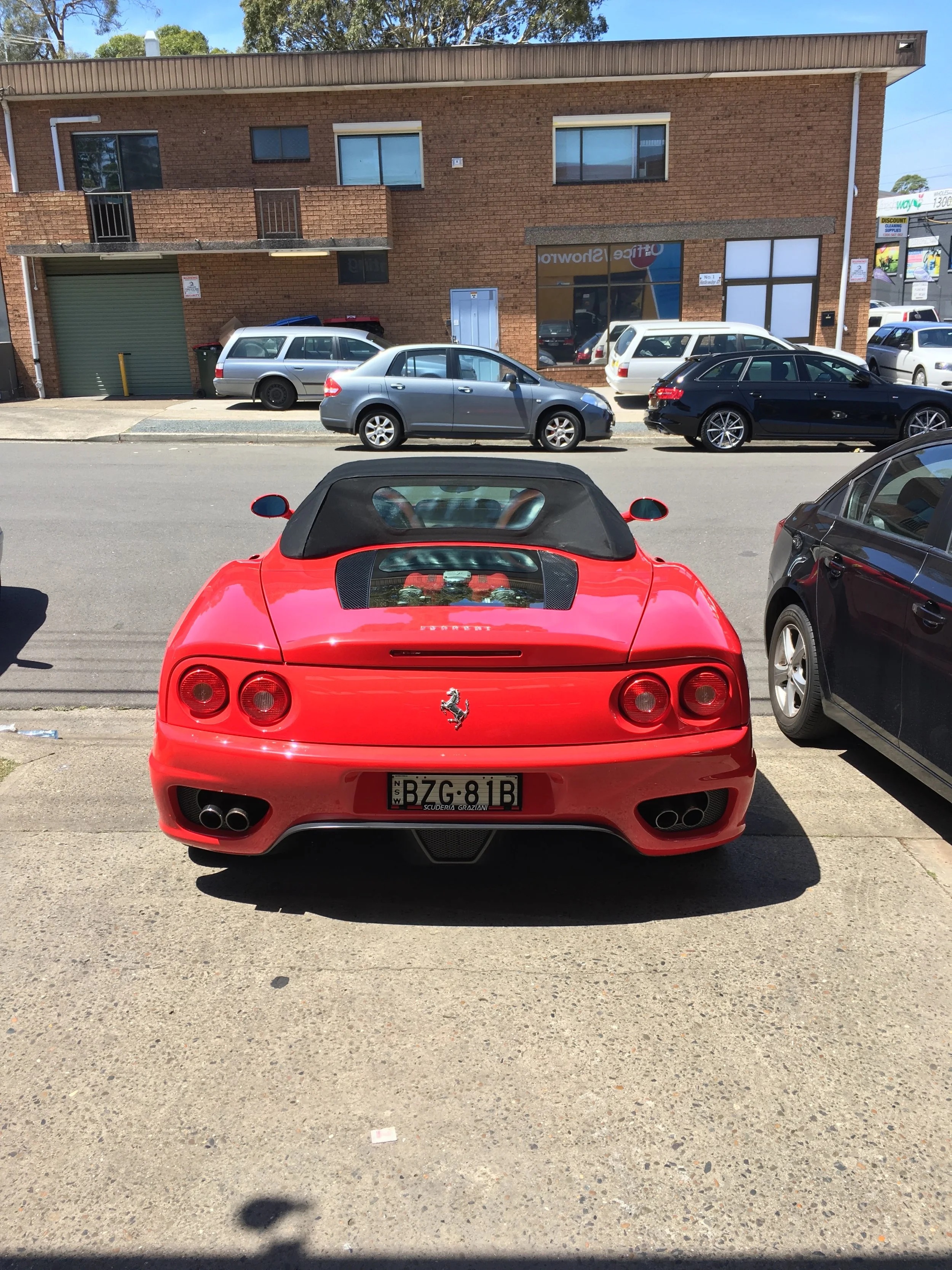 Red Ferrari sports car viewed from the back, parked on a street with other cars and a brick building in the background.