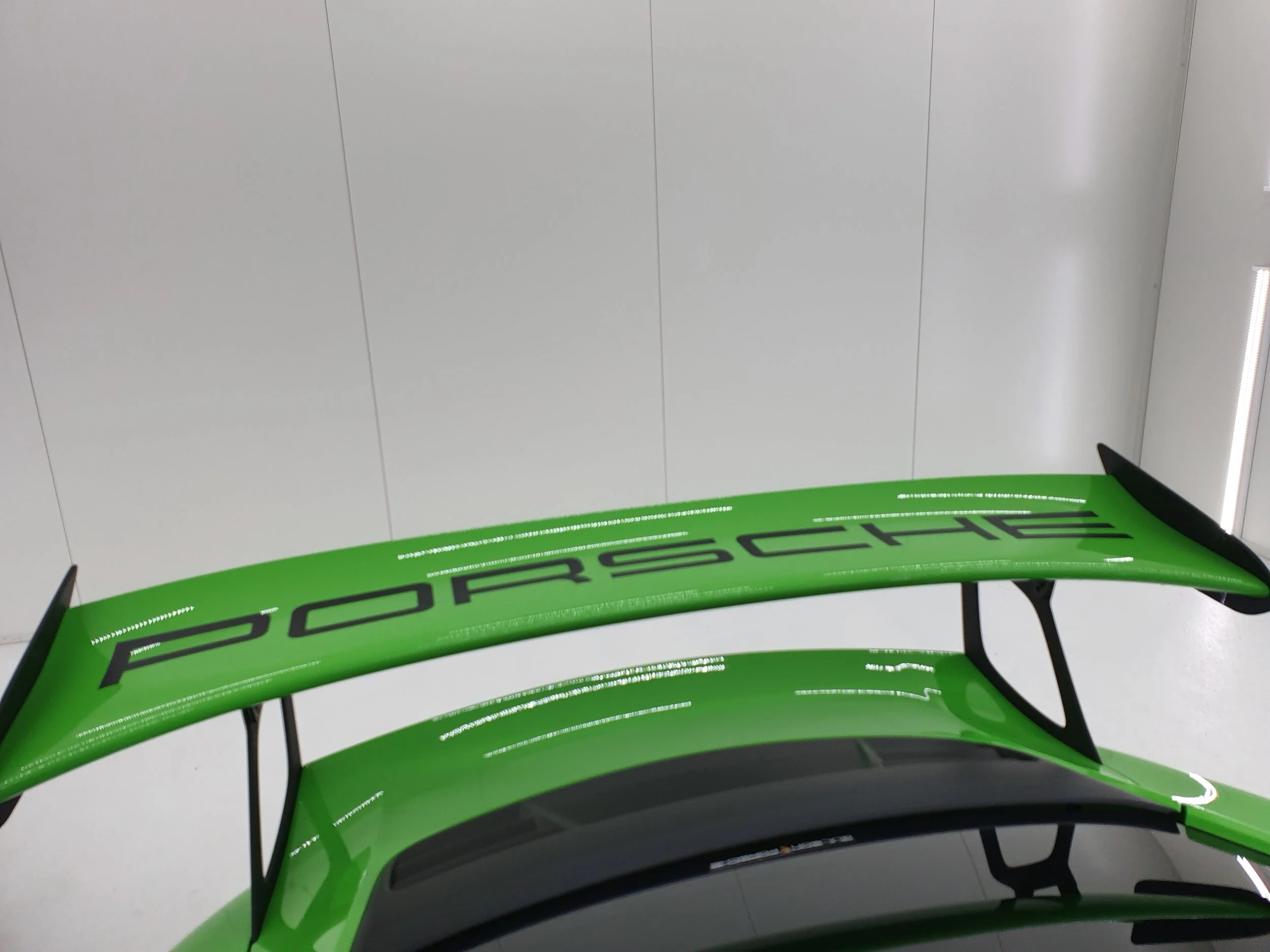 The image shows the rear wing of a bright green Porsche race car with the Porsche logo and branding visible on it, placed in an indoor setting with plain white walls and ceiling lights.