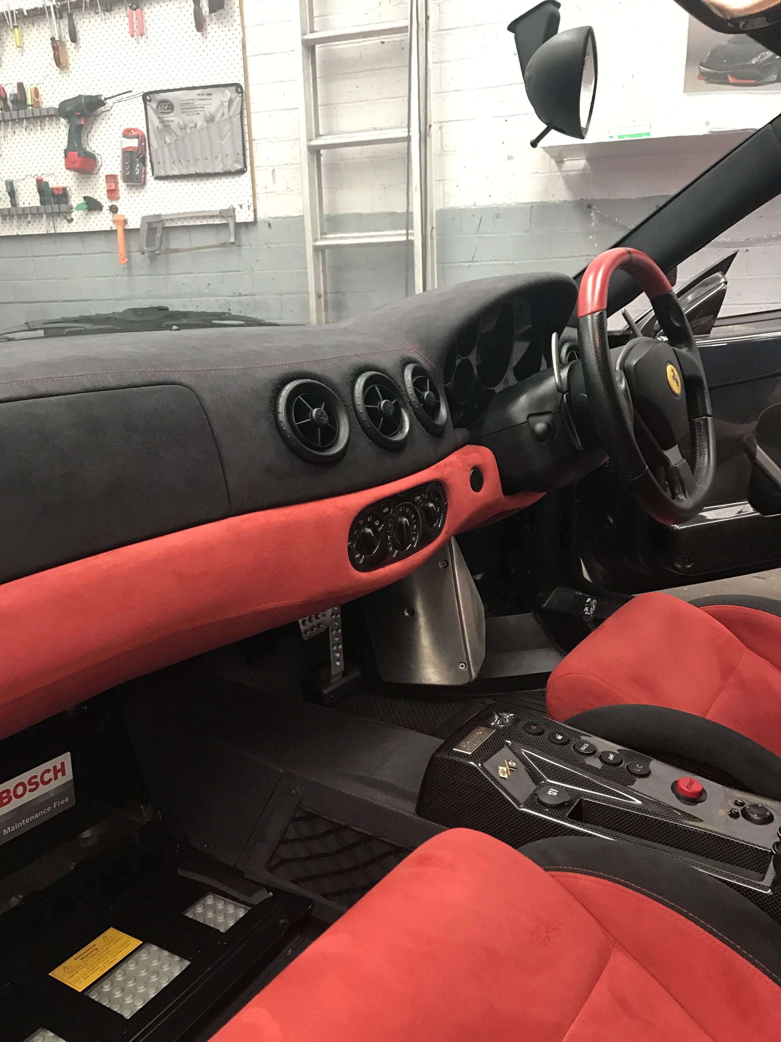 The interior of a sports car with a red and black leather steering wheel, red seat, and a dashboard with three round air vents and various controls. The background shows a garage with tools hanging on a pegboard and a ladder against a white brick wal