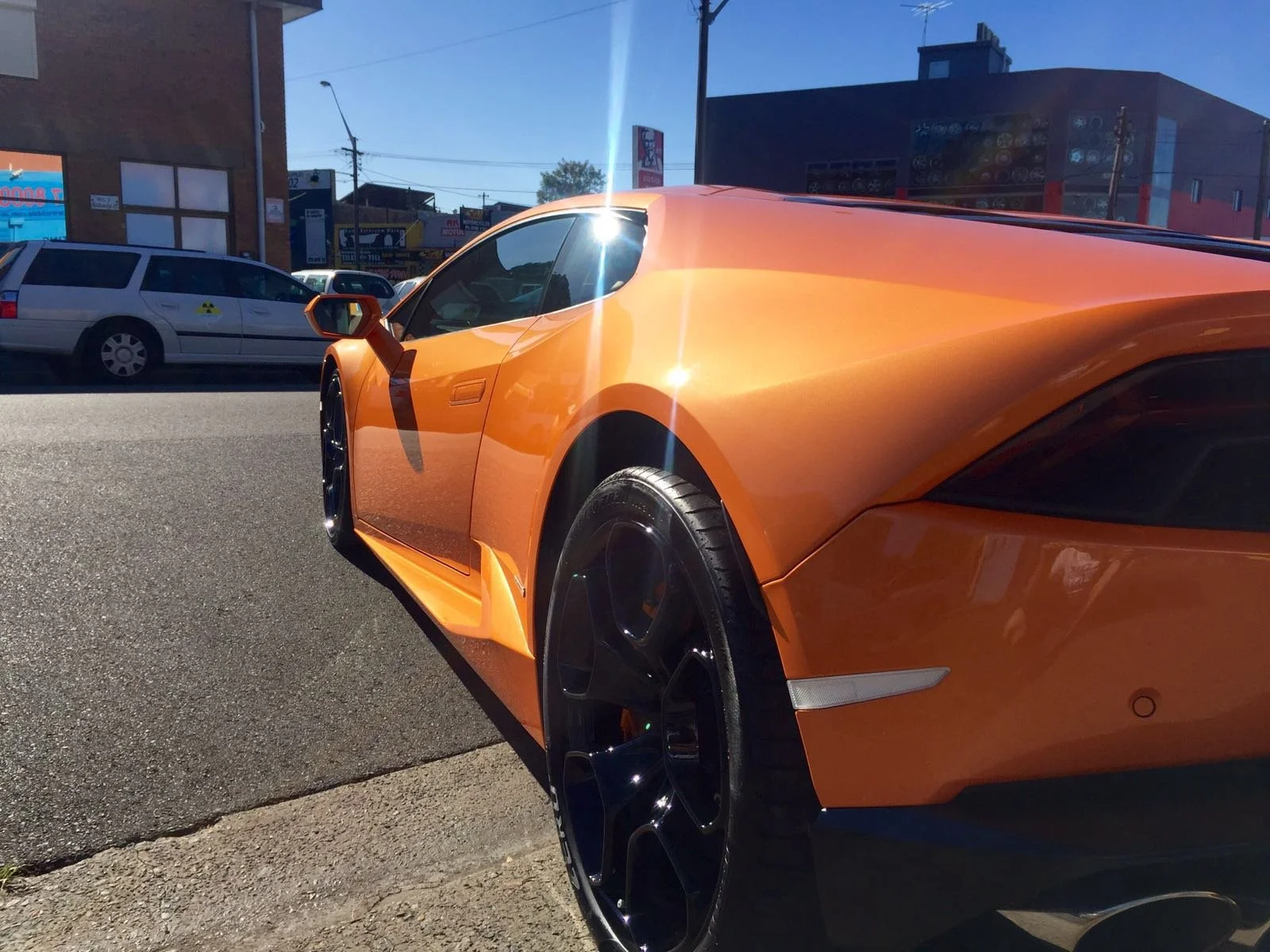 Close-up of an orange Lamborghini parked on a city street with buildings and a blue sky in the background.
